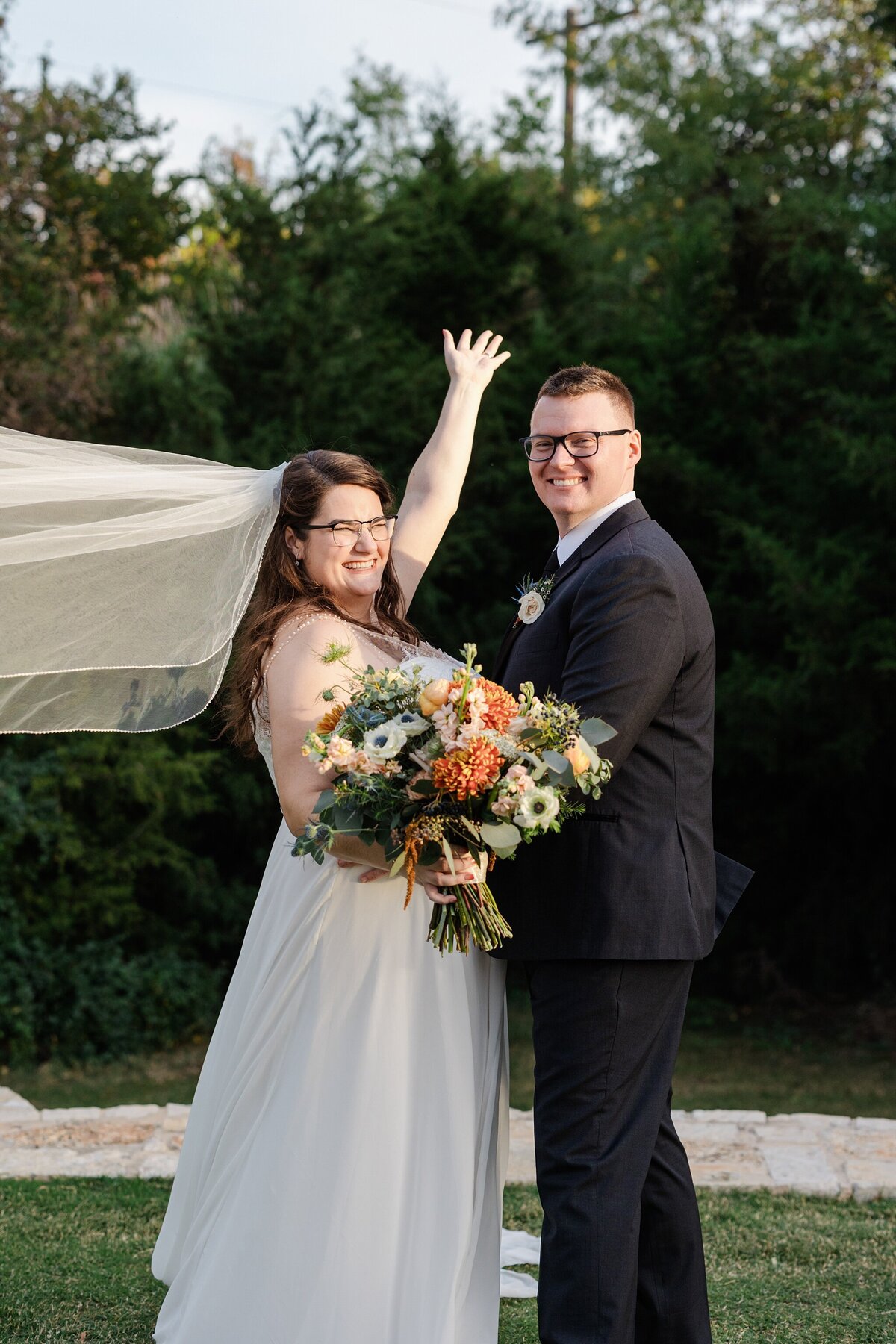 A portrait of a bride and groom posing outside after their wedding ceremony at The Laurel in Grapevine, Texas. The bride is on the left and is wearing a long flowing, sleeveless, white dress with a long veil flowing out behind her while holding a large, colorful bouquet. The groom is on the right and is waring a dark suit with a boutonniere.