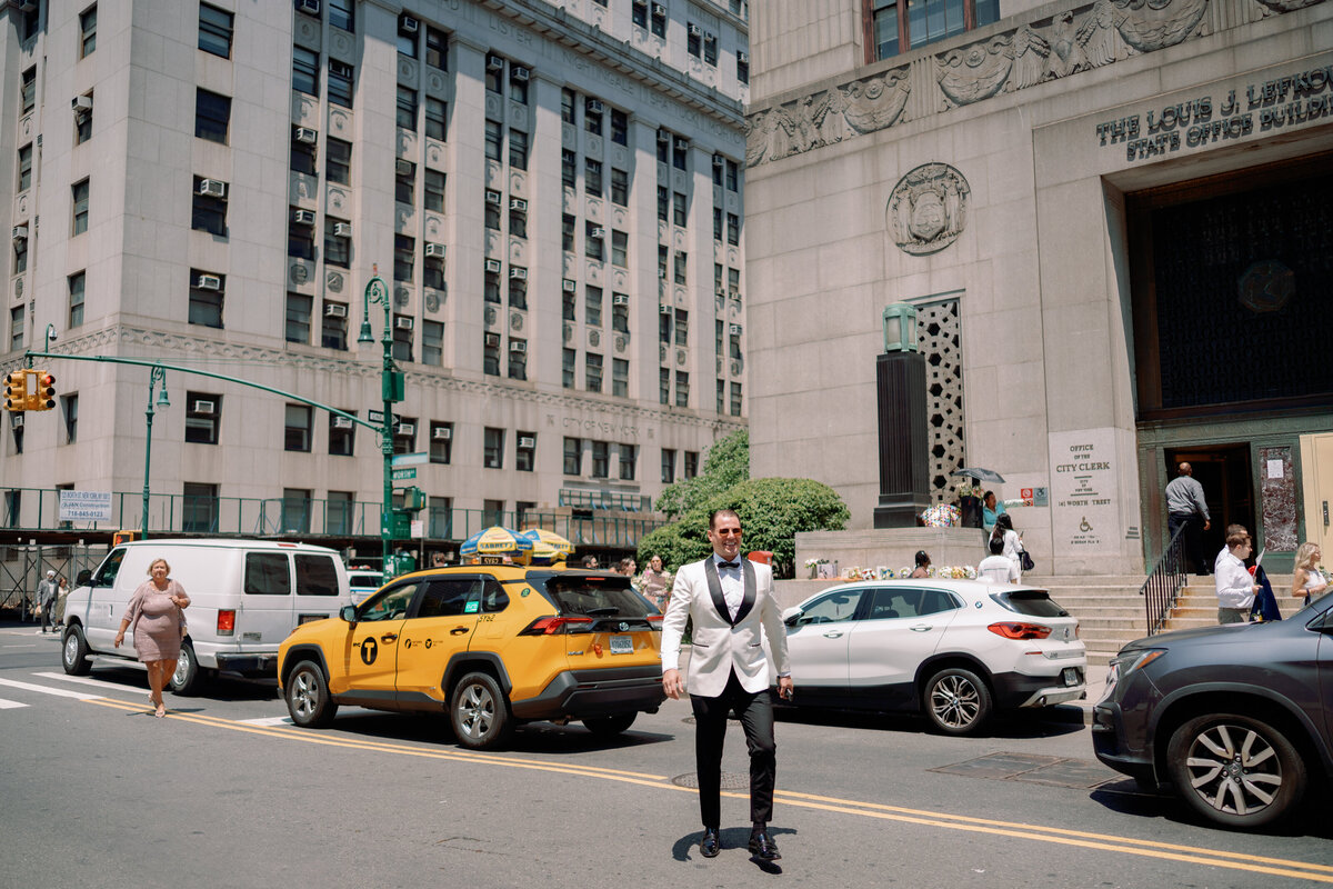 Groom in a white tuxedo jacket standing on the streets of New York City with yellow taxis and historic buildings in the background, captured during Japna and Chris’s Hotel Chelsea elopement by NYC wedding photographer Perry Hancock.
