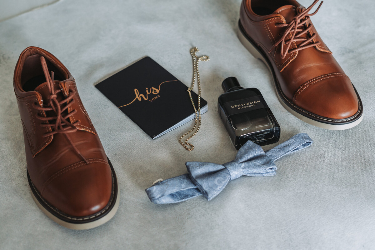 lat lay of groom’s wedding accessories including shoes, bow tie, vow book, and cologne at Lesner Inn, Virginia Beach, Virginia.