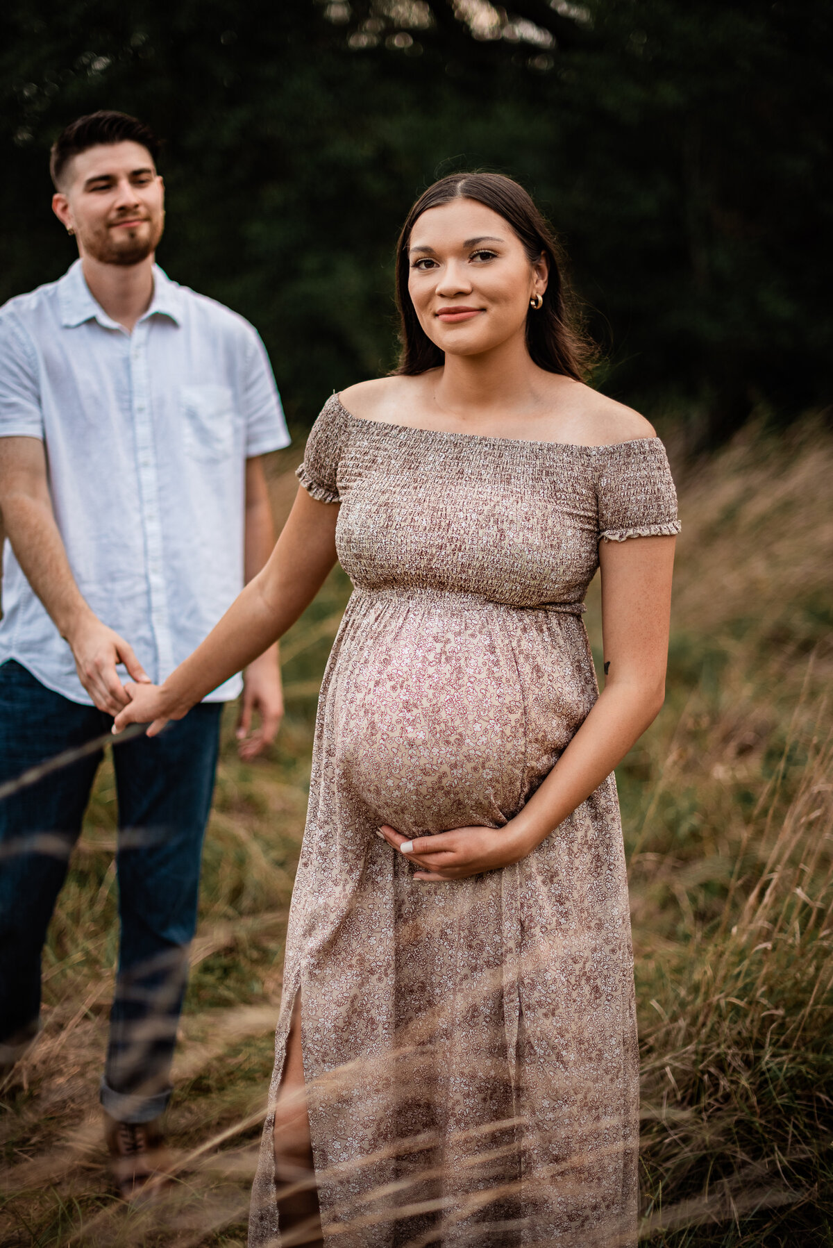 A pregnant woman holds her boyfriend's hand and smiles at the camera while he looks at her lovingly.