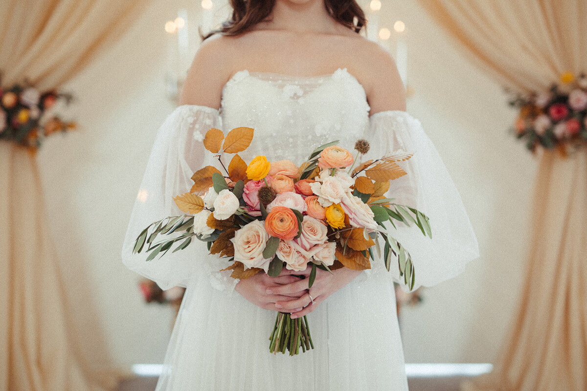 A close-up of bride wearing a white wedding gown holding a bouquet of orange, yellow and blush-colored flowers.