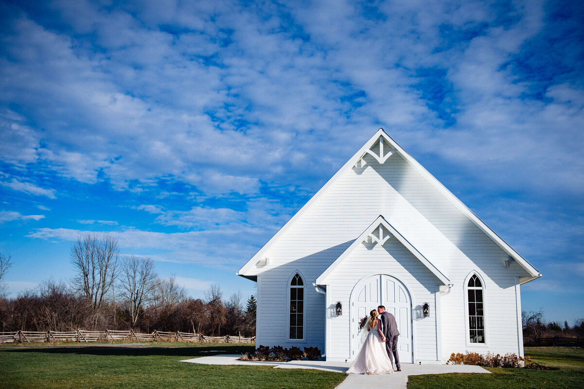 a scenic photo with a big blue sky showing a bride and groom kissing in front of the Stonefields Estate wedding venue Ceremony House.  Captured by Ottawa Wedding Photographer JEMMAN Photography
