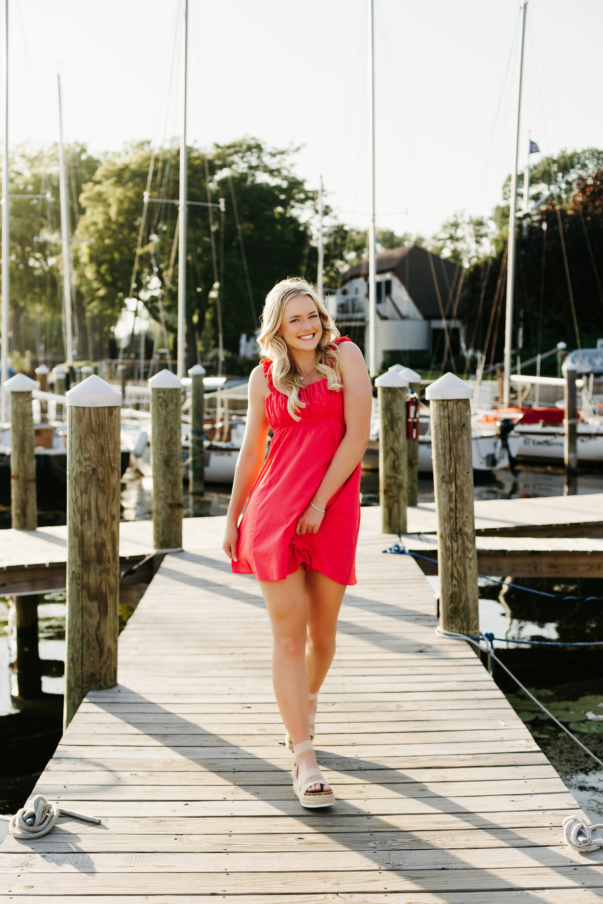 Senior girl in a red dress walking down a dock with sailboats in the background