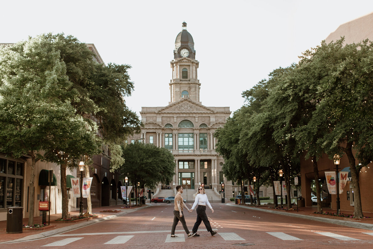 A cute and candid moment of an engaged couple walking in Sundance Square with the Tarrant County Courthouse in the background. Captured by Fort Worth Wedding Photographer, Megan Christine Studio