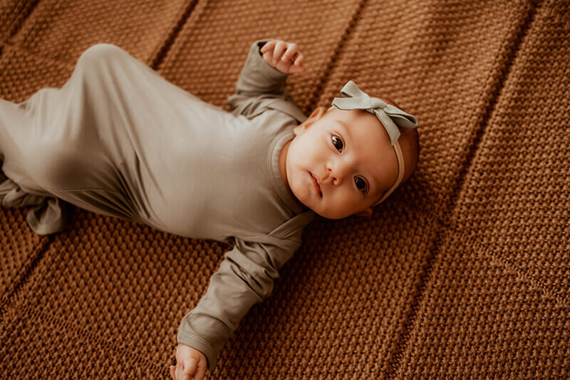 newborn baby girl laying on a brown knit blanket while wearing an olive green sleeper and bow