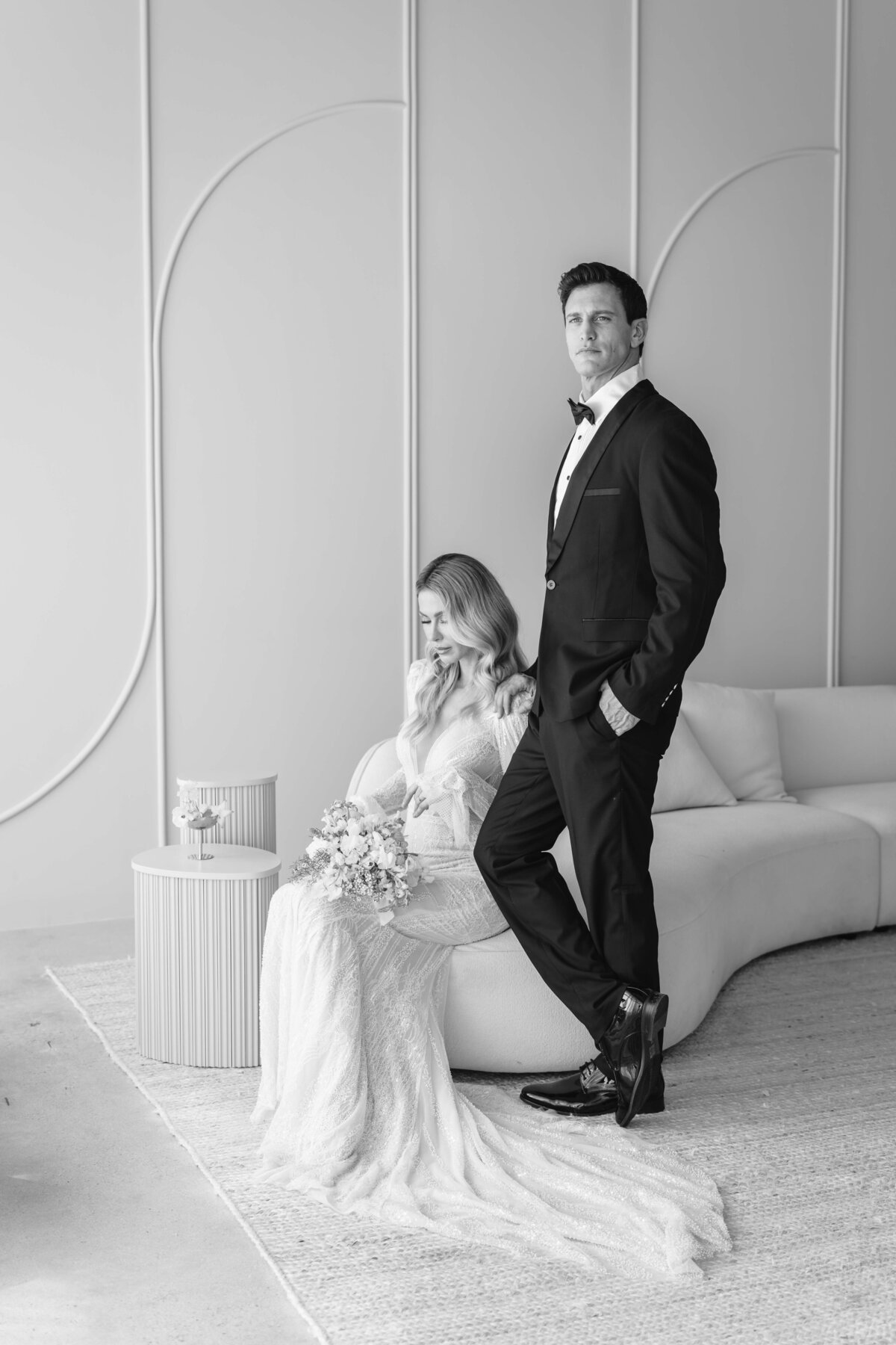 black and white of a bride sitting looking at her bouquete, and the groom standing nest to her