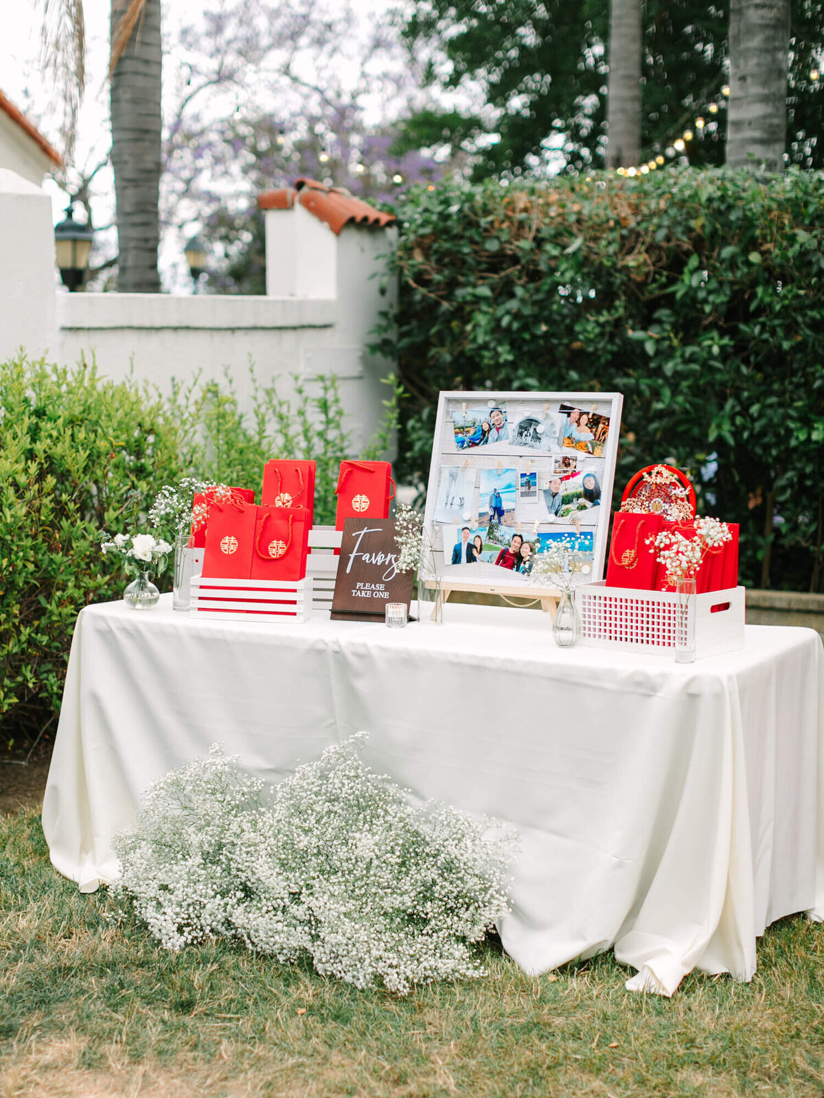 Outdoor table with red gift bags and a collage of photos on display. White tablecloth, surrounded by greenery and white flowers. Festive and welcoming mood.