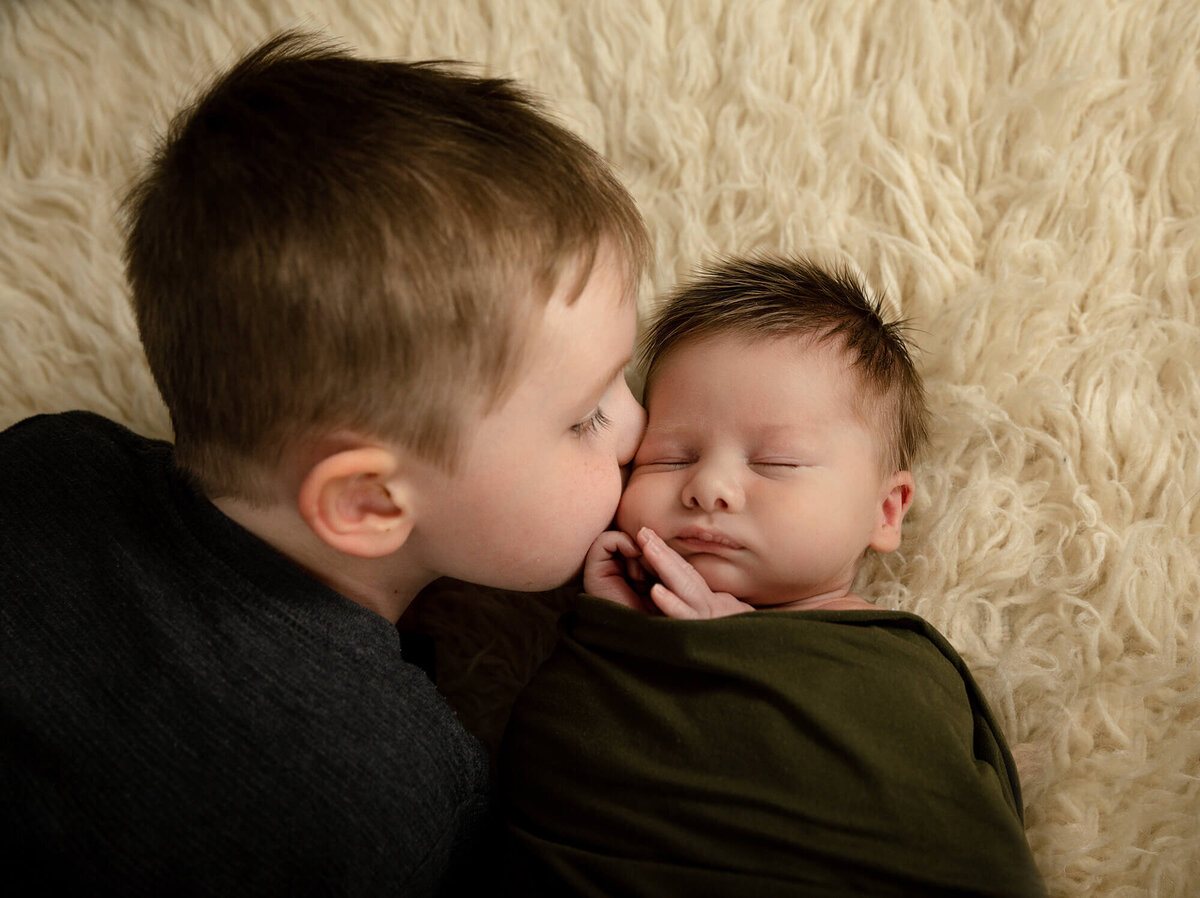 Older brother looking at his newborn brother laying on a creme colored rug.