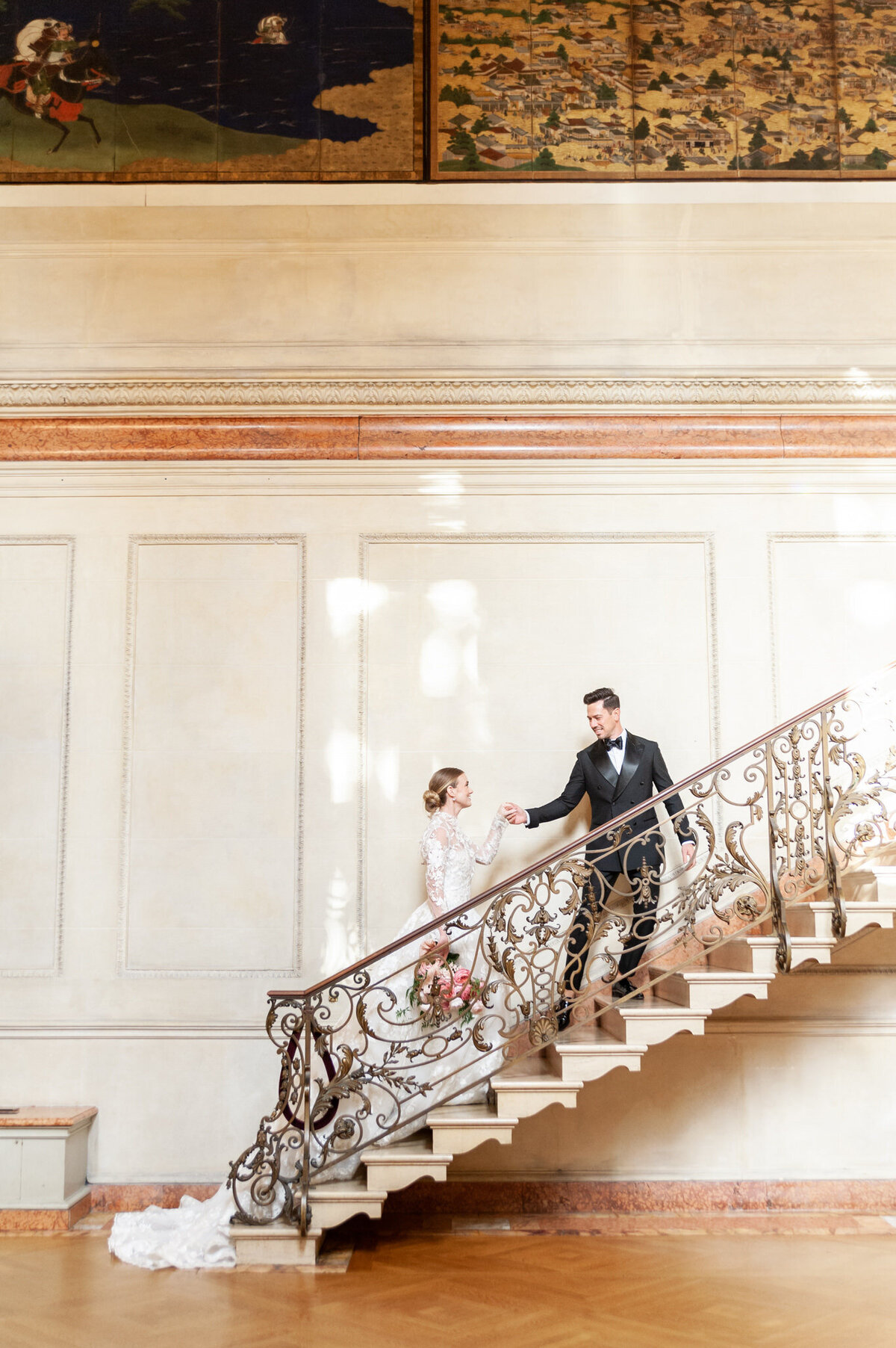 groom helping his bride up the marble stairs in a mansion
