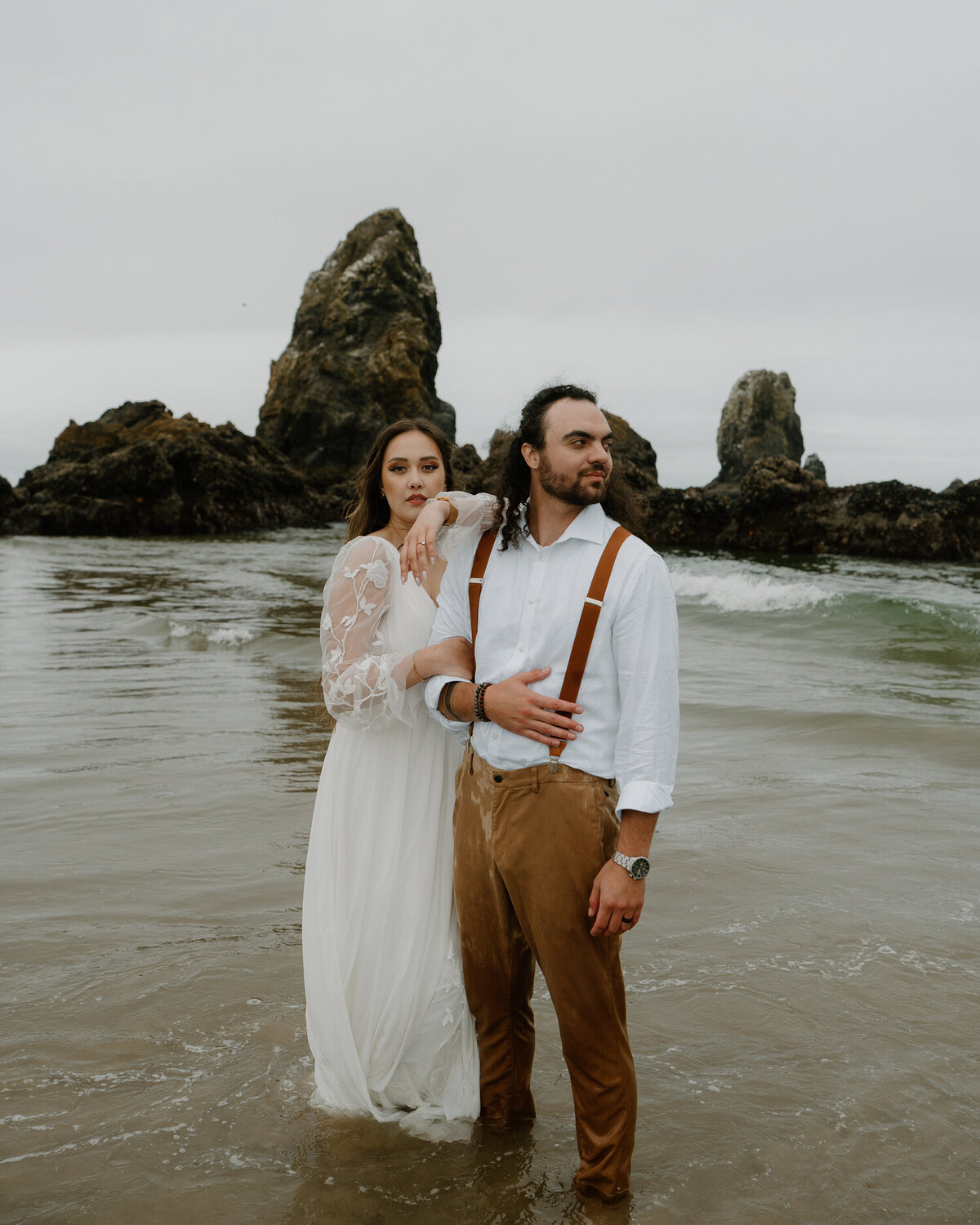 A couple stands in the ocean in front of black rocks at their Cannon Beach elopement. 