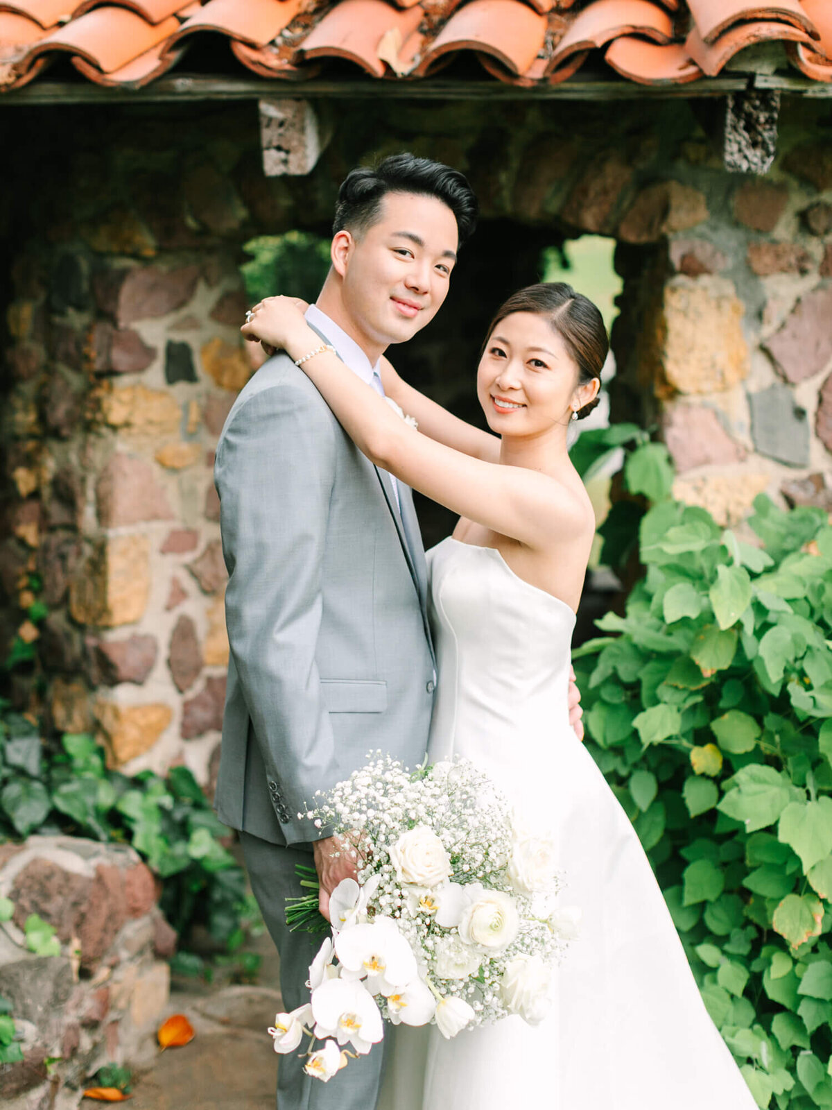 A bride and groom pose lovingly by a rustic stone archway. The bride holds a bouquet of white flowers, wearing a strapless gown. The groom is in a light gray suit. Lush greenery surrounds them, creating a serene and romantic atmosphere.