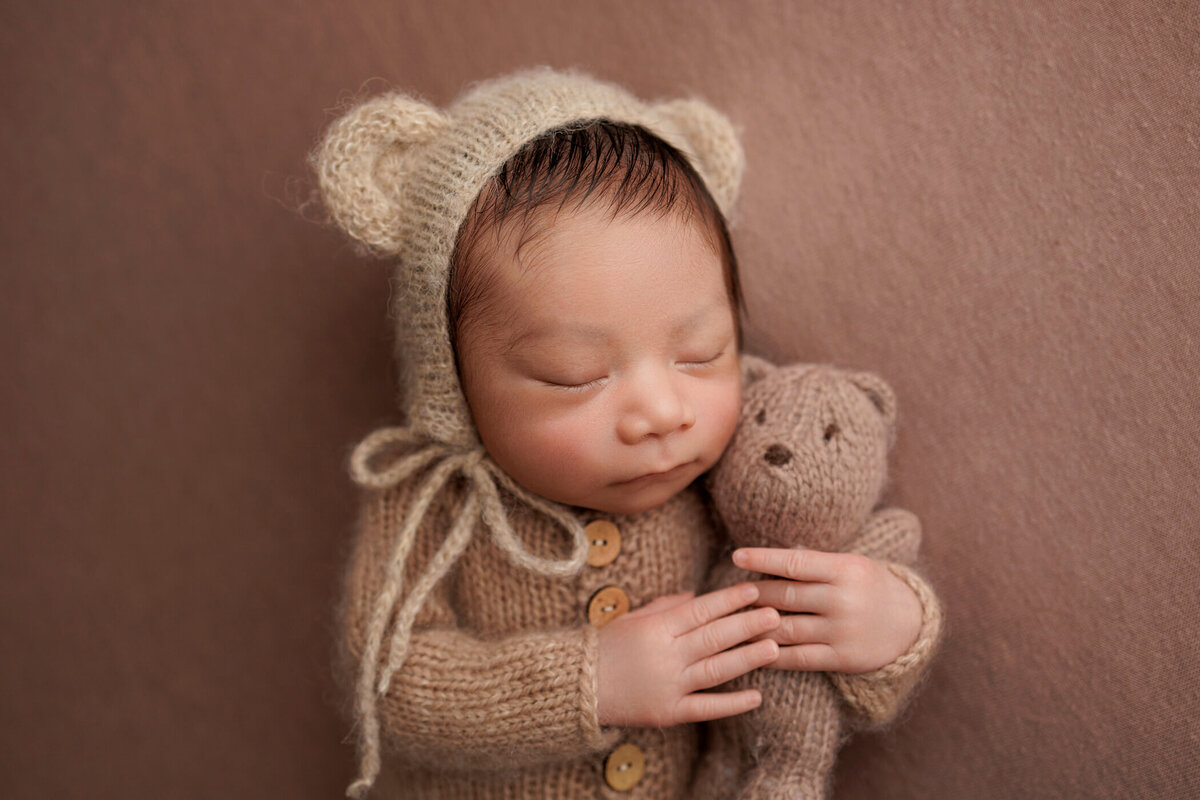 Newborn baby wearing a knitted bear costume, sleeping and holding a small knitted teddy bear on a brown background.