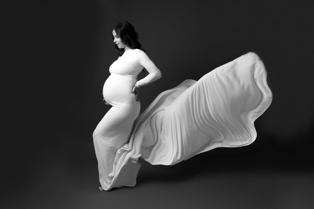 A maternity portrait of a woman wearing a white gown with a long flowy tail posing side ways showing her pregnant belly