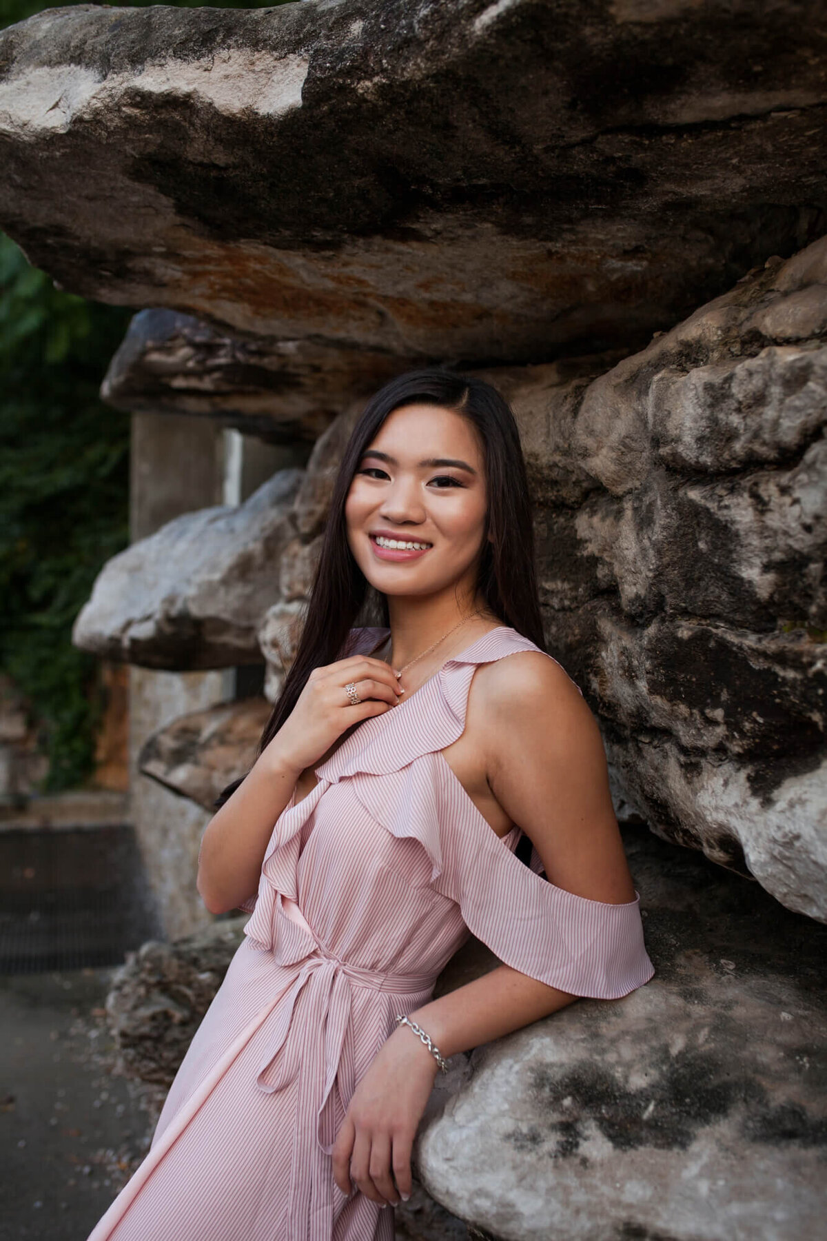 A lovely Asian American teen wearing a cold shoulder light pink dress leans against a rock wall. Captured by Springfield, MO senior photographer Dynae Levingston.