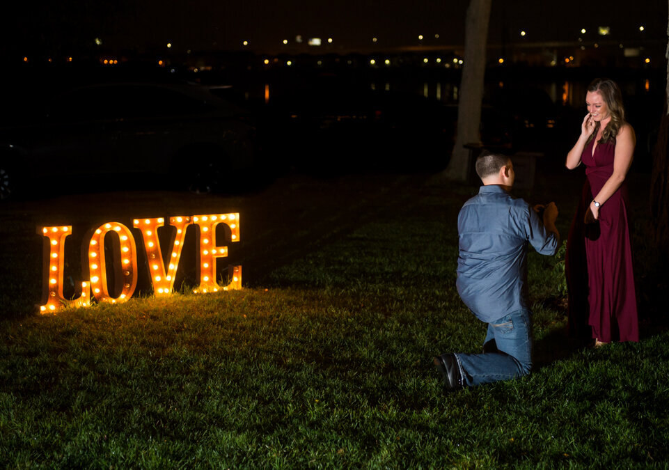 A man kneels down and proposes in front of a lit up love sign.