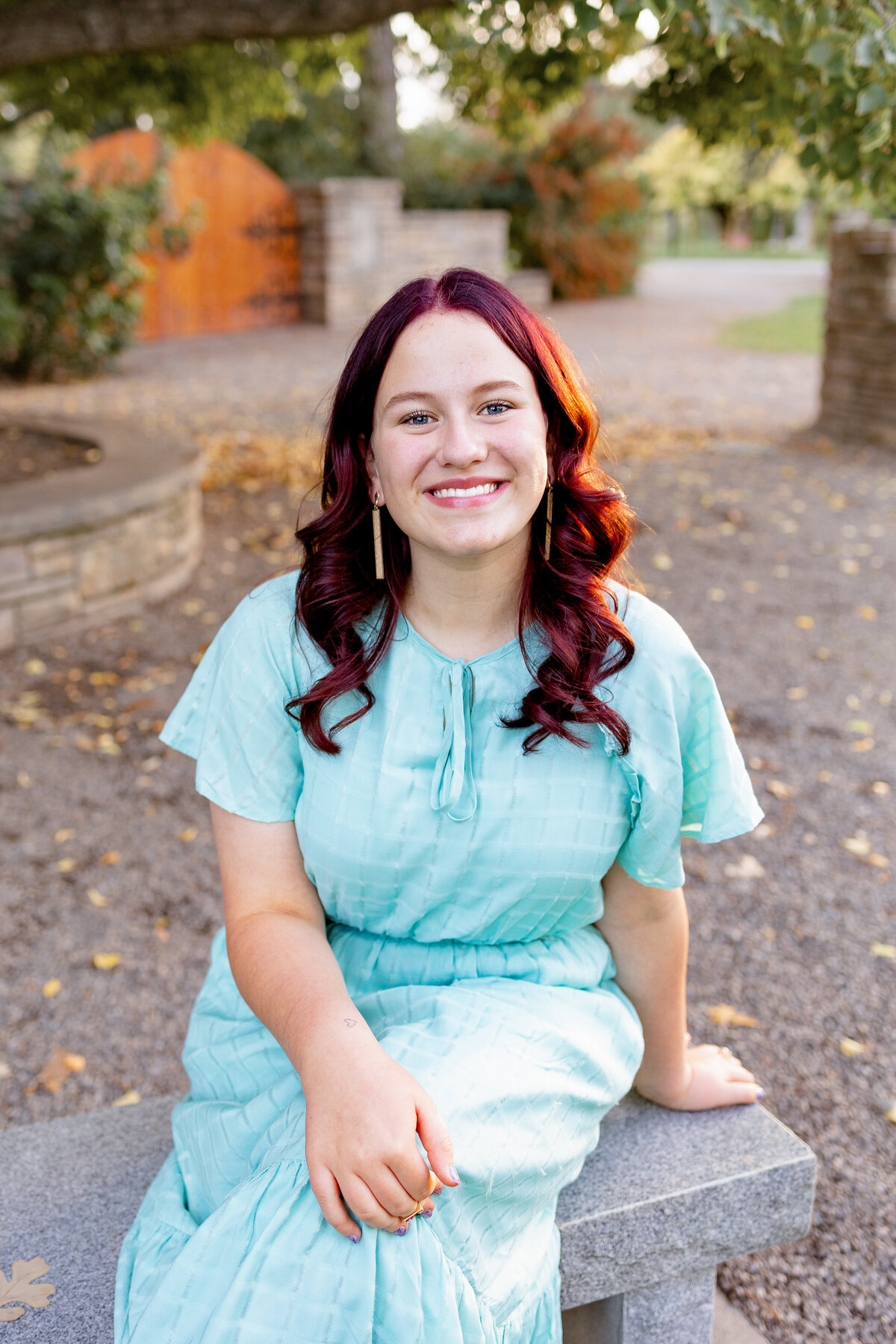 A young woman with red hair sits on a stone bench in a garden setting, smiling at the camera. She wears a light blue dress, and the background features a wooden gate, stone walls, and lush greenery.