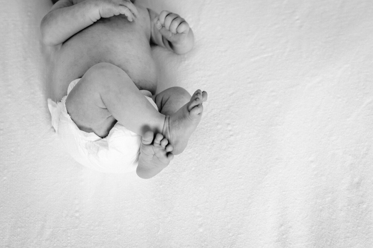 A black-and-white image focused on the legs and diapered bottom of a newborn baby lying on a soft white surface, with the baby's feet crossed at the ankles.
