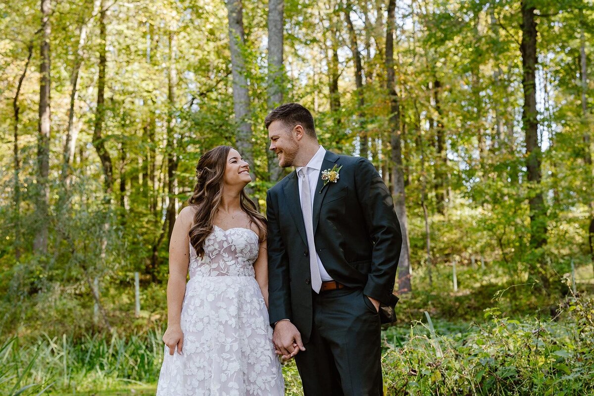 A bride and groom holding hands and smiling at one another on their wedding day.  They are standing in the woods. 