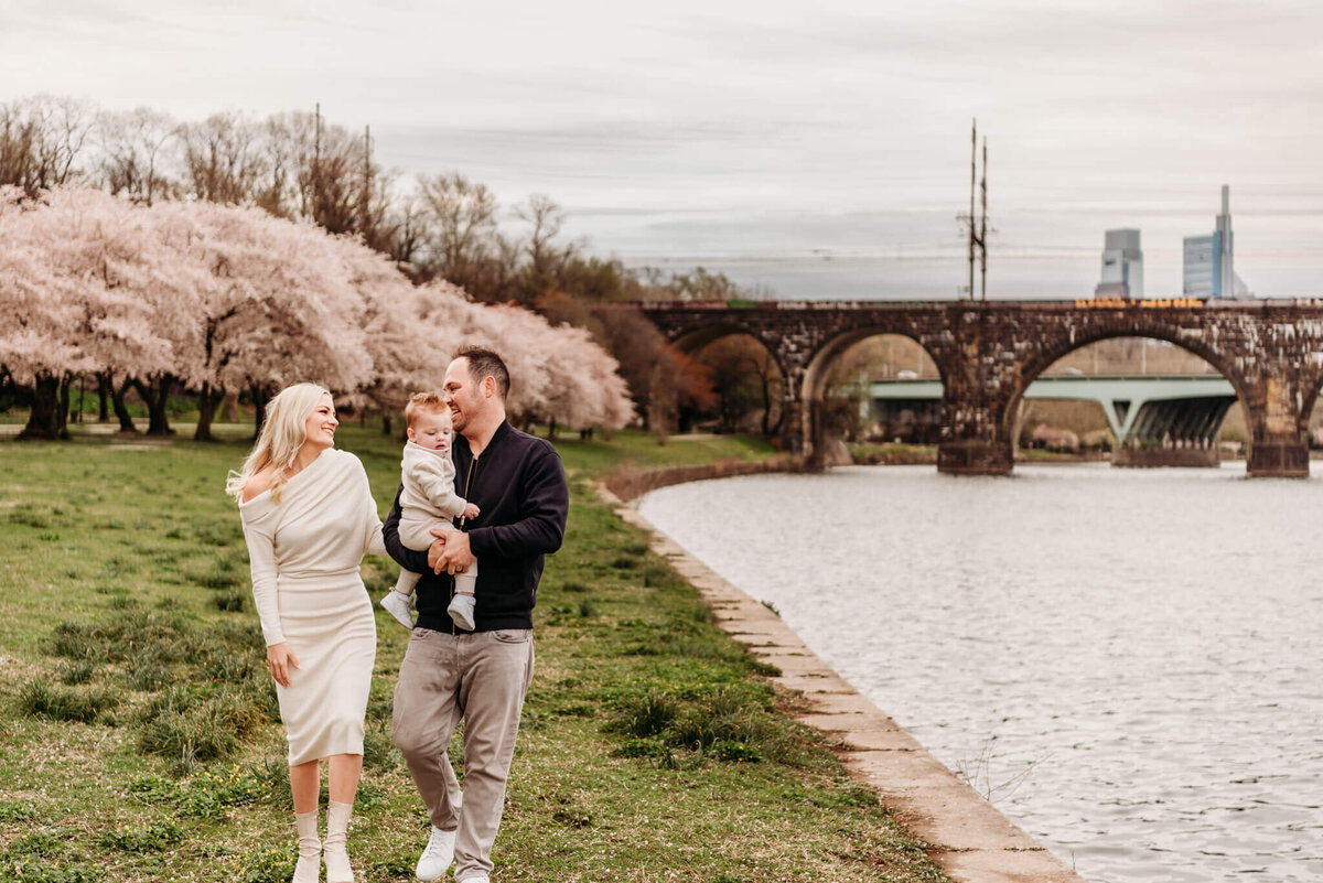 A man is holding one year old son while walking next to his wife along the Schyukill River with Cherry Blossom trees blossoming in the background.  Photo taken during their Philadelphia family photography session.