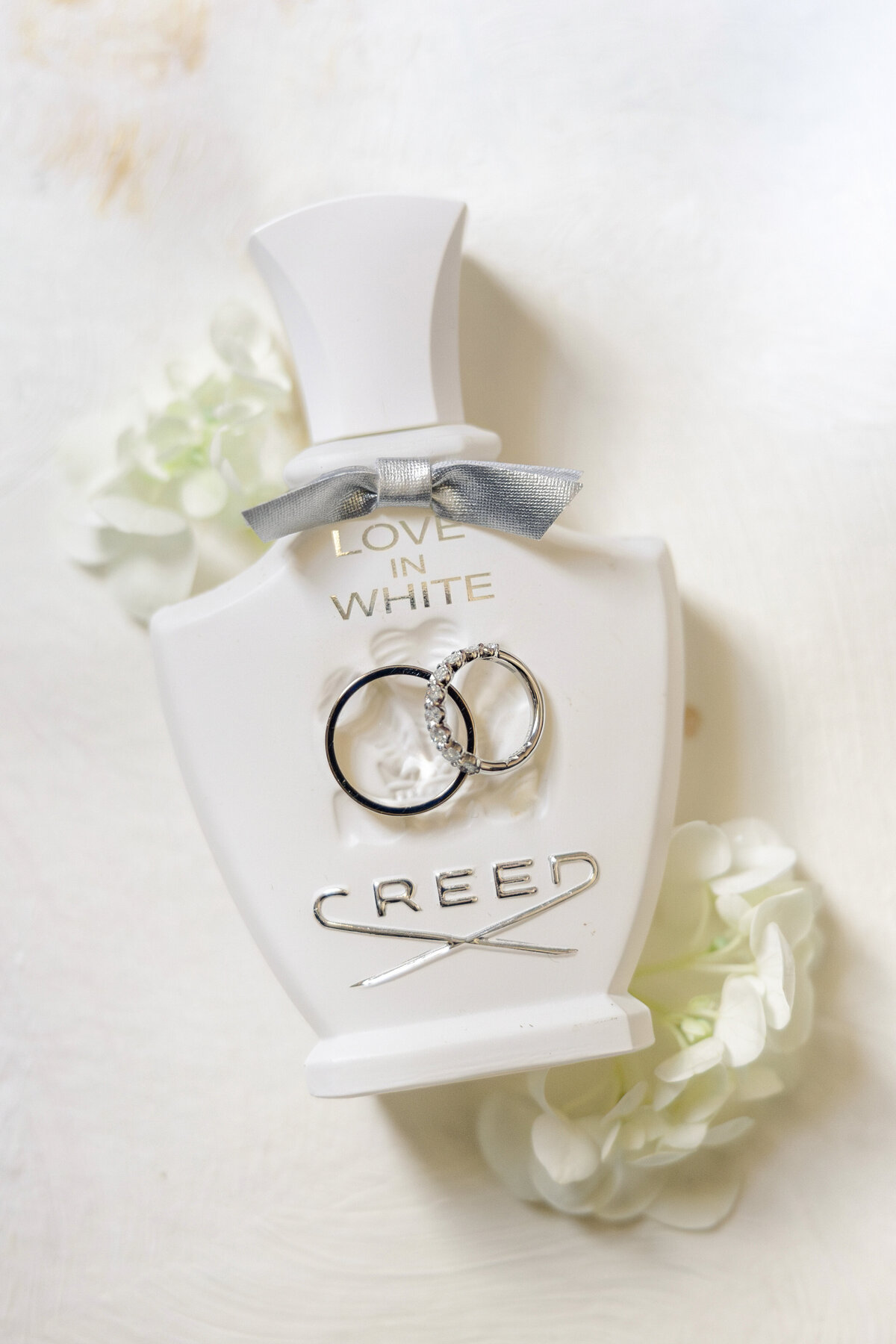 A white Creed perfume bottle labeled Love in White is adorned with two silver rings, one featuring a diamond. The bottle is topped with a gray bow, and white flowers are softly blurred around it on a light background.