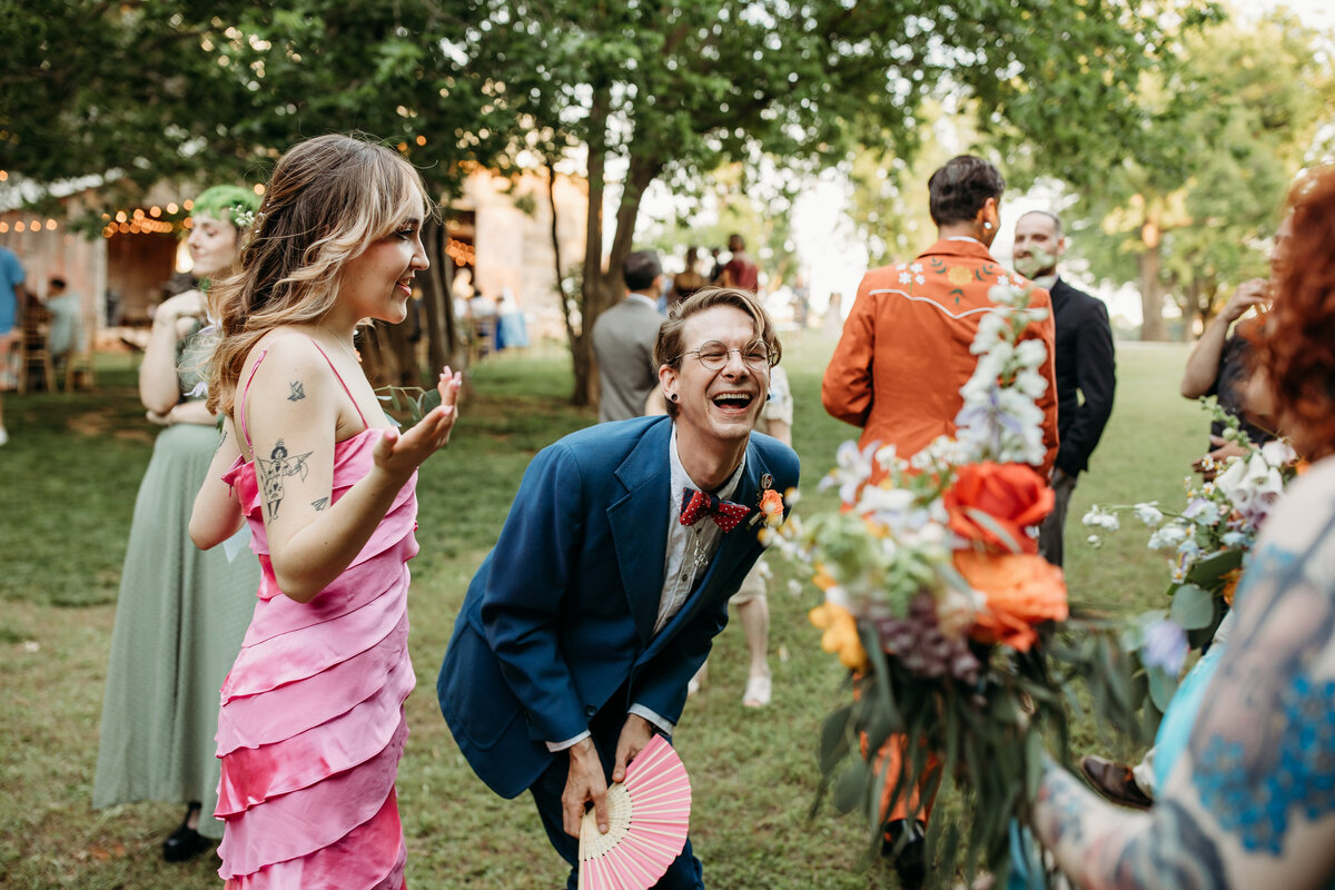 Joyous wedding guests laughing and dancing outdoors, with a man in a blue suit and woman in a pink dress in focus