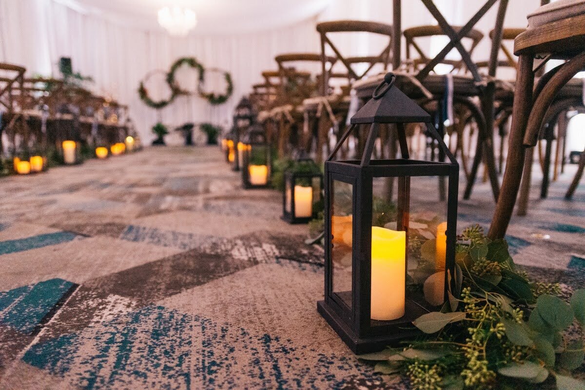 Candle in a lantern and eucalyptus greens line the aisle of an indoor wedding ceremony.