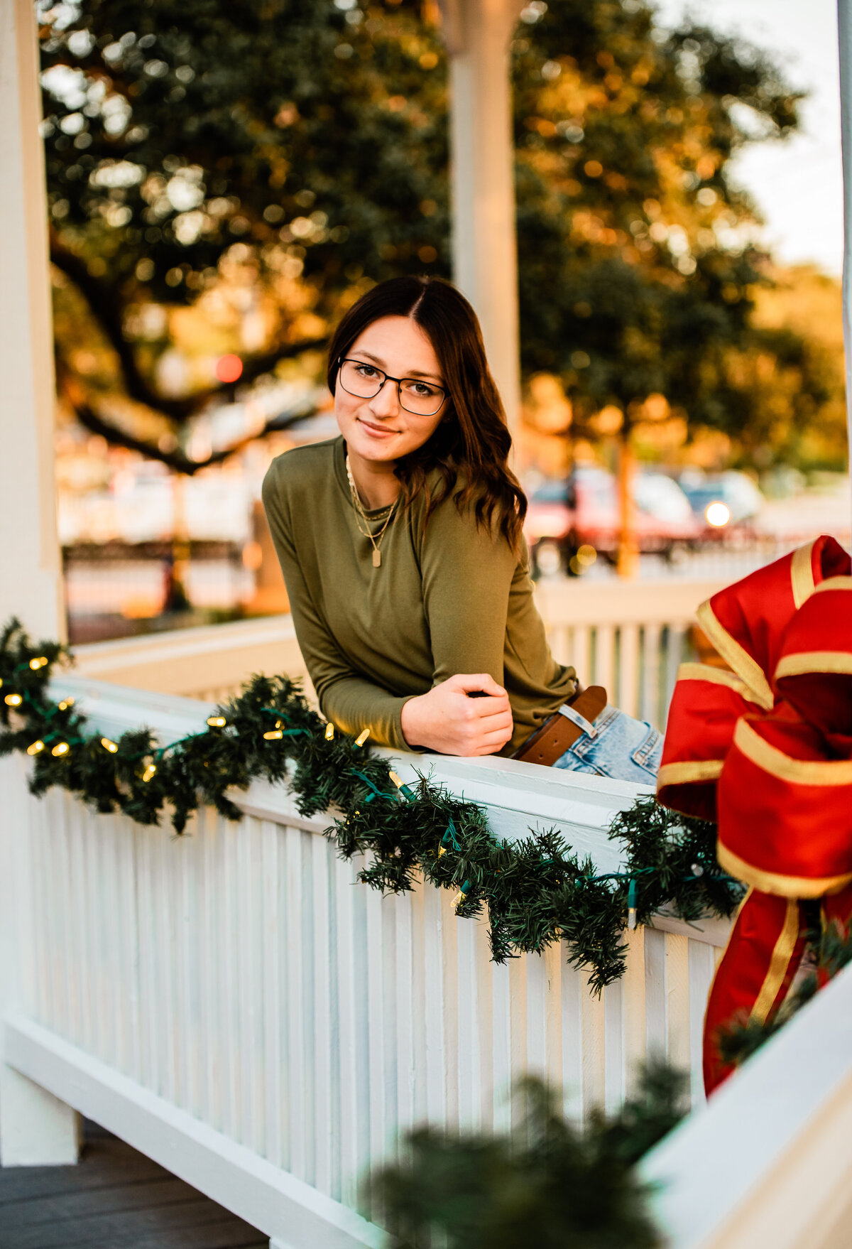 A senior girl leans over a white gazebo railing that is decorated with Christmas garland,