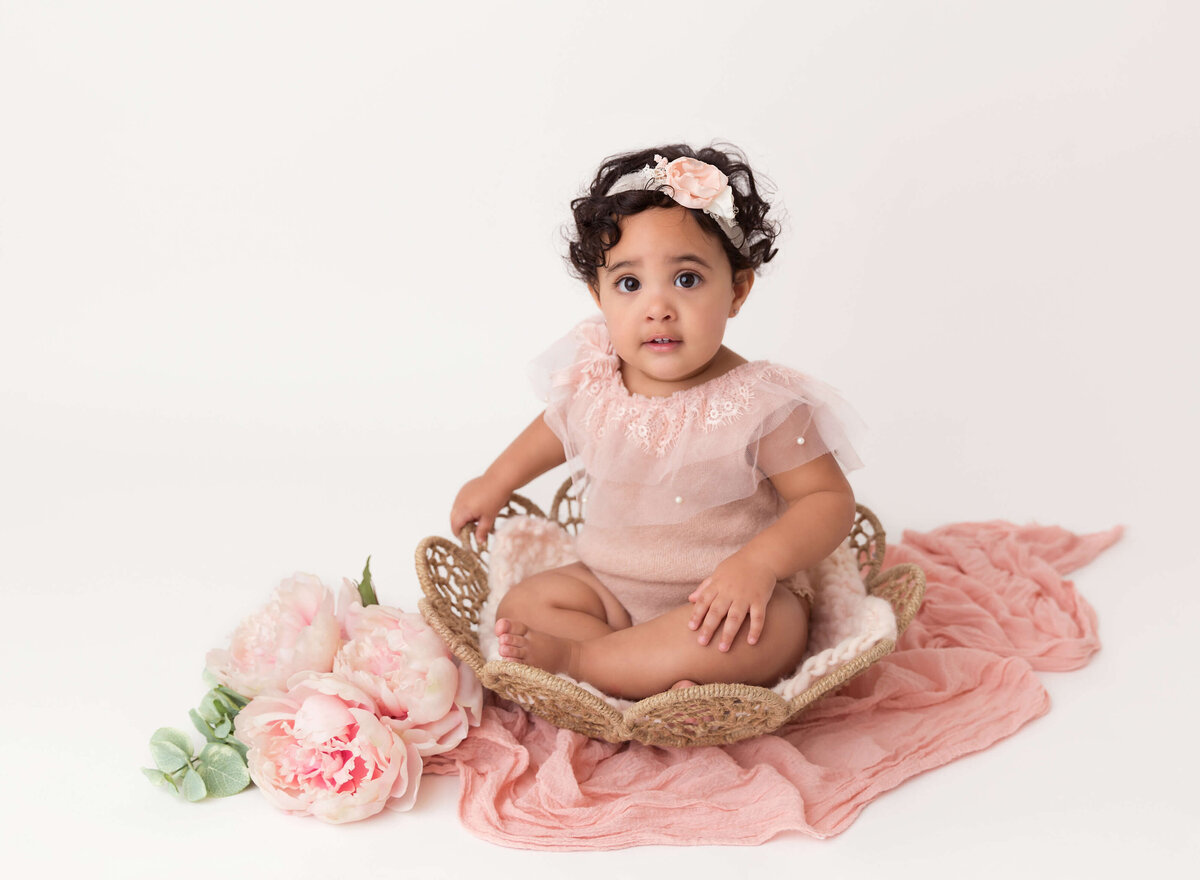 6-month-old baby girl in a blush onesie with organza details sitting in a rattan basket for her milestone photoshoot in NYC
