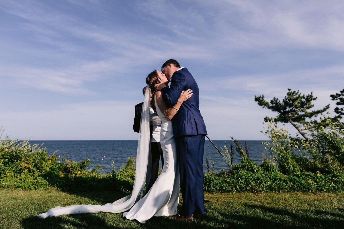 The groom is kissing the bride in a wedding ceremony at Wianno, Cape Cod, MA, with the sea and sky in the background.