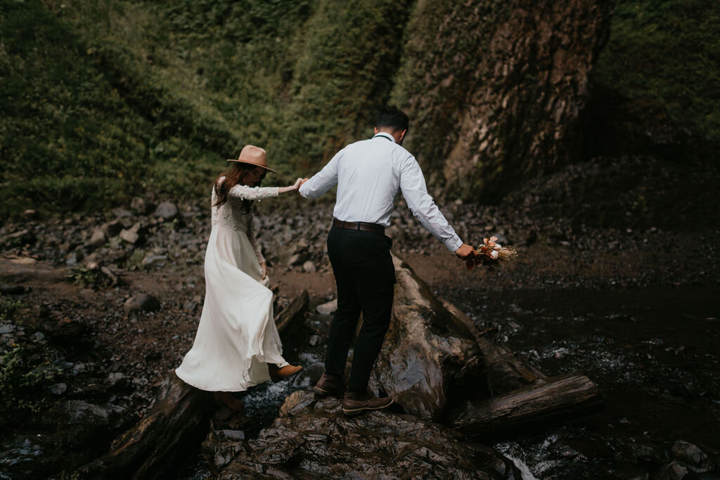 Bride and groom helping each other cross a river on their way to their waterfall elopement