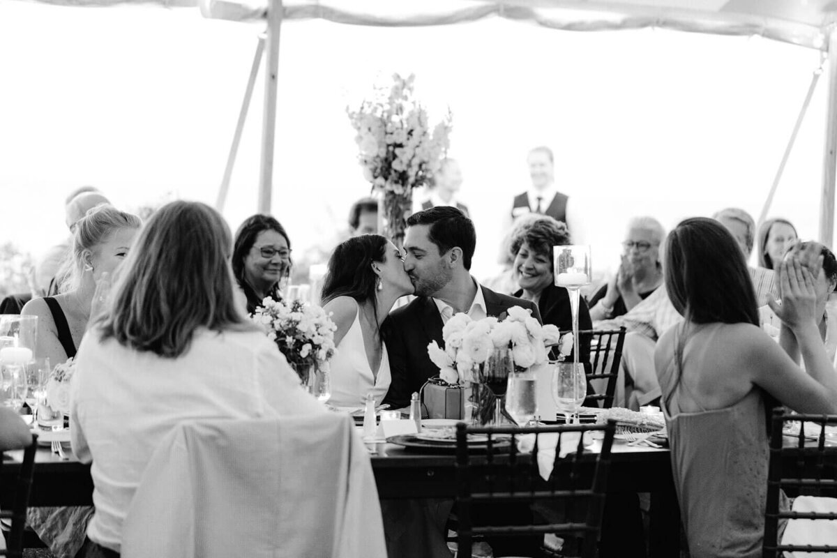 Black and white photo of the bride and the groom kissing as guests cheer on, in Cape Cod Summer Tent, MA.