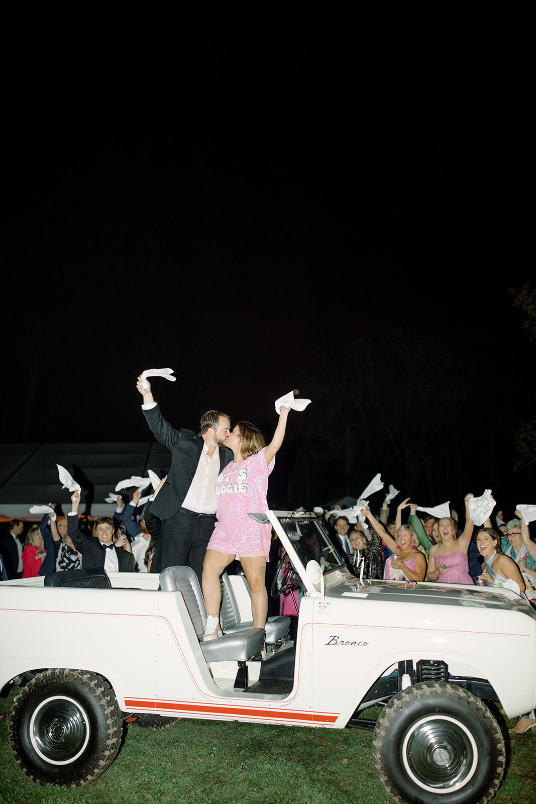 Couple standing on a white Bronco at their wedding reception exit while wedding guests wave napkins and cheer