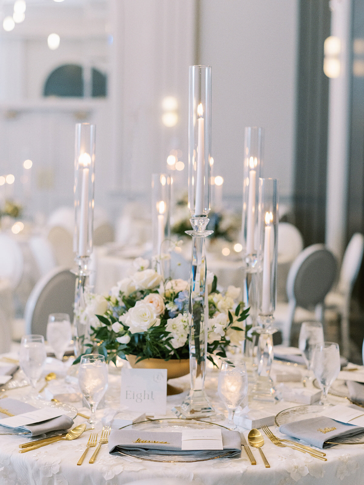 A round table set for an event by a Banff wedding planner features tall candle holders, a floral centerpiece, and place settings with plates, gold cutlery, and menus. A card with the number eight sits on the table.