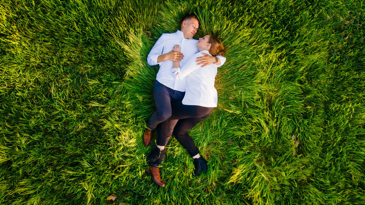 Engaged couple wearing white shirts and black pants lay in the grass cuddling in each other's arms during an engagement session. Drone captures the photo from the air.  Taken by philippe studio pro, a wedding photographer studio in Sacramento.