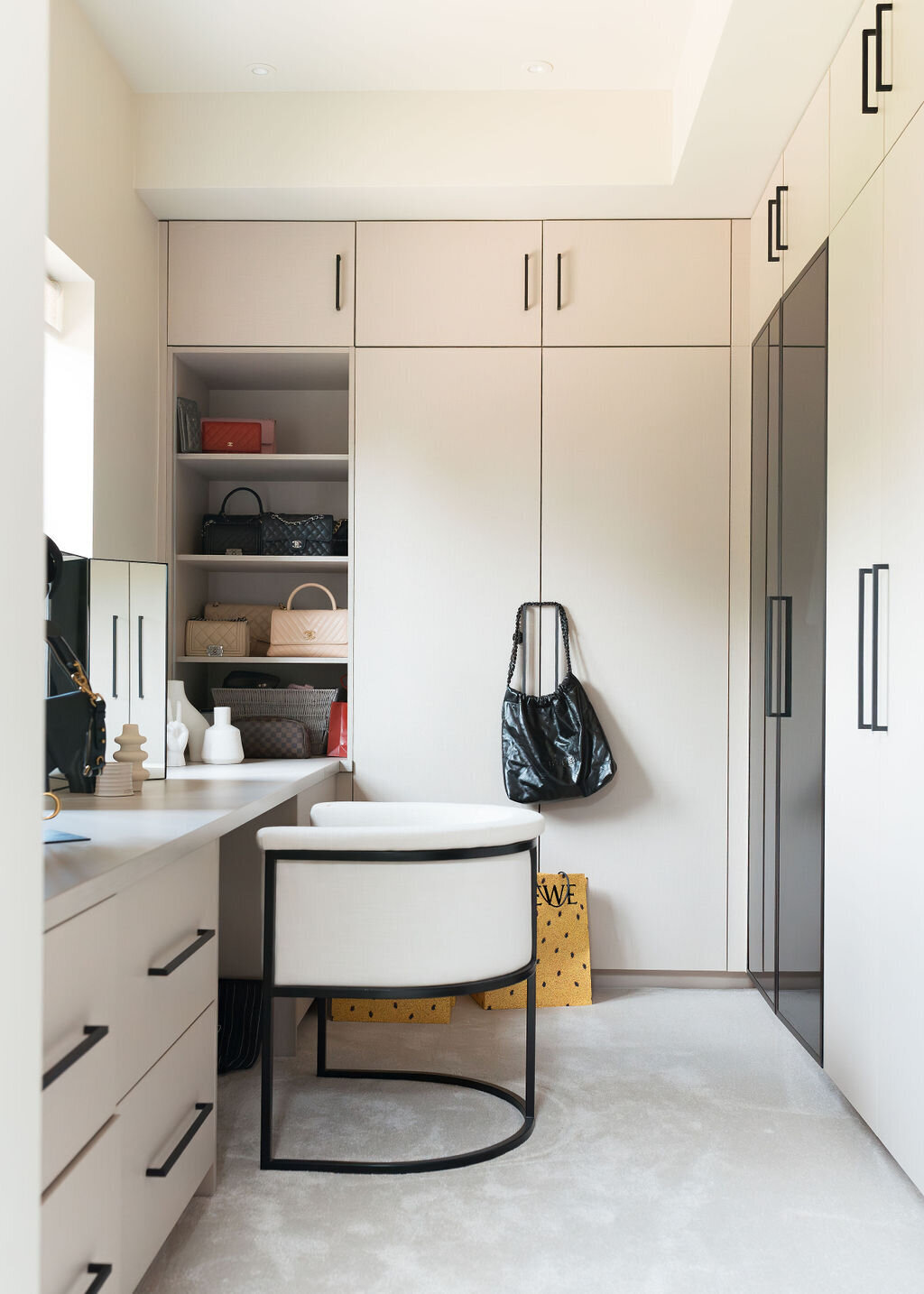 A modern, light-filled walk-in closet with white cabinets and built-in shelves containing handbags and storage baskets. A white desk with a black-framed chair is in the corner, and the floor is carpeted in light neutral.