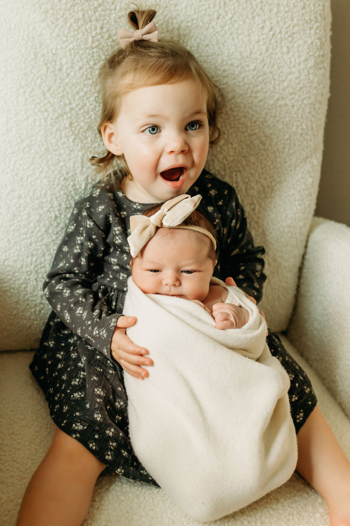 A toddler girl sits in a rocking chair inside a nursery holding her newborn baby sister