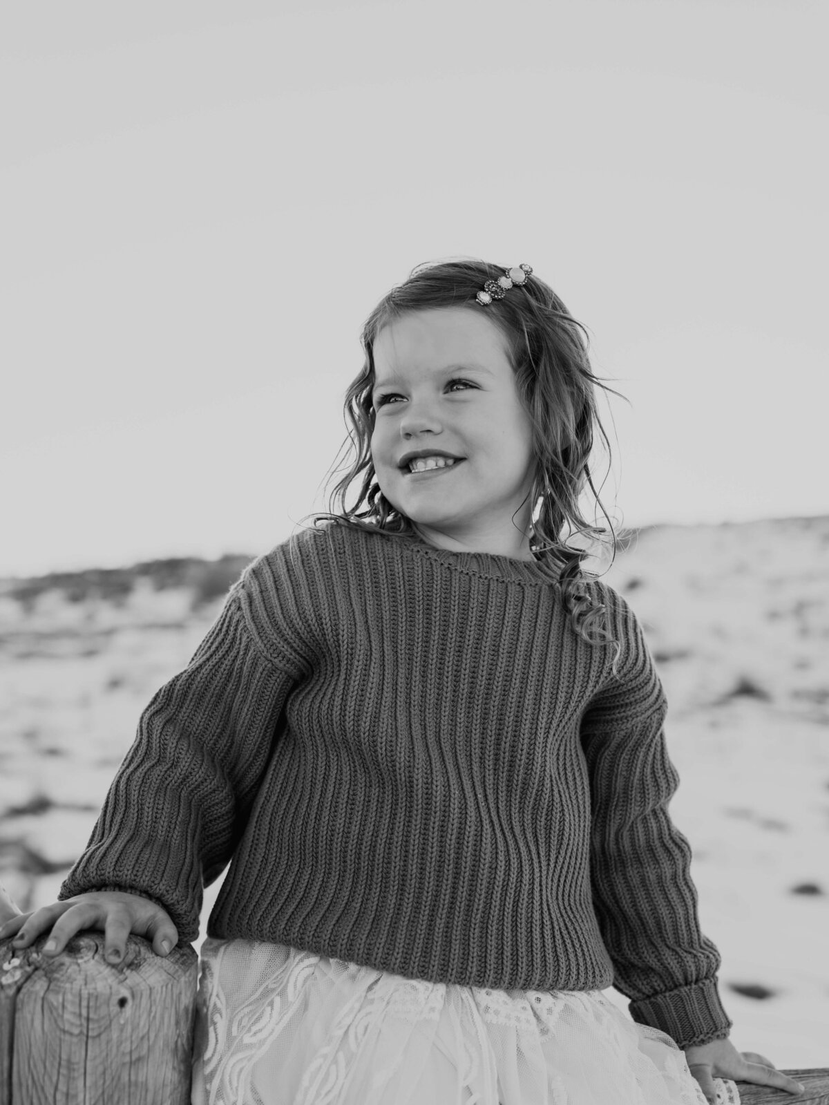 A black and white close-up portrait of a young toddler, smiling as she looks off into the distance with a snowy backdrop.