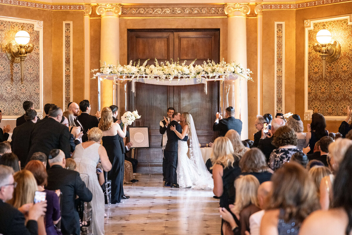 A bride and groom share a kiss under a floral arch during their wedding ceremony in an elegant, ornate room. Guests are seated, some standing, capturing the moment on phones. Clergy members are nearby, and the atmosphere is joyful and celebratory.