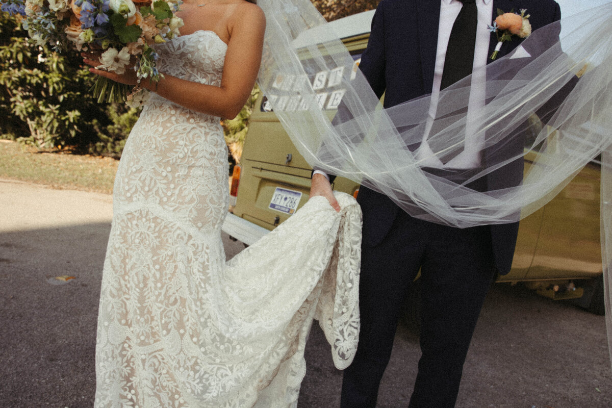 Groom holding the train and veil of his bride's wedding dress in front of a VW Bus