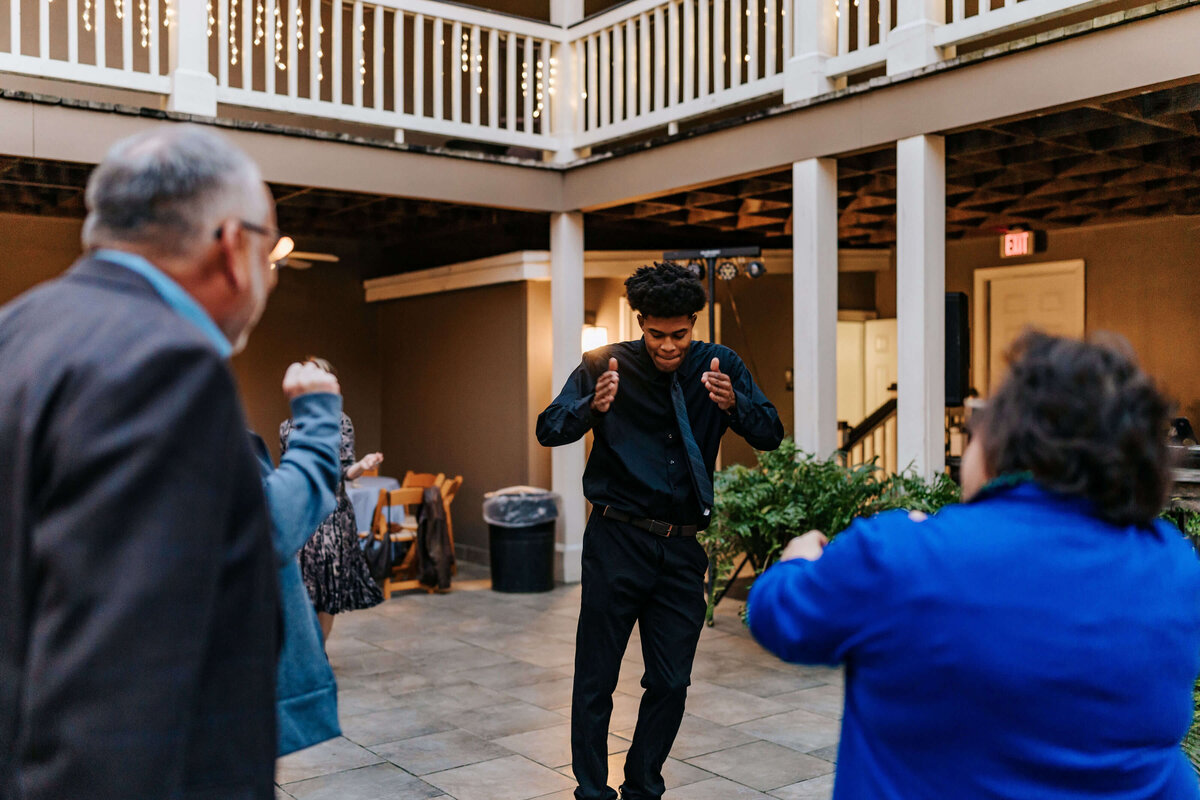 A young man in black dances with a smile in a courtyard, while two older adults in blue jackets face him, enjoying the moment. String lights and a balcony are visible above.