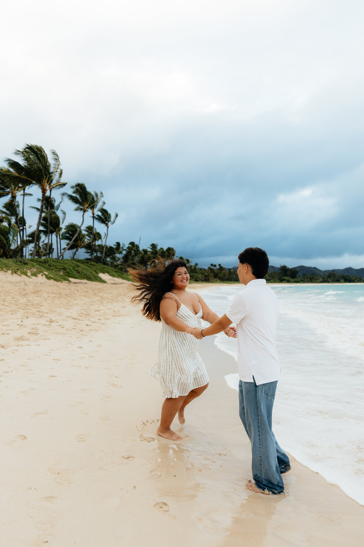 Couple laughing and dancing together barefoot on the beach in Oʻahu with palm trees and ocean waves in the background.