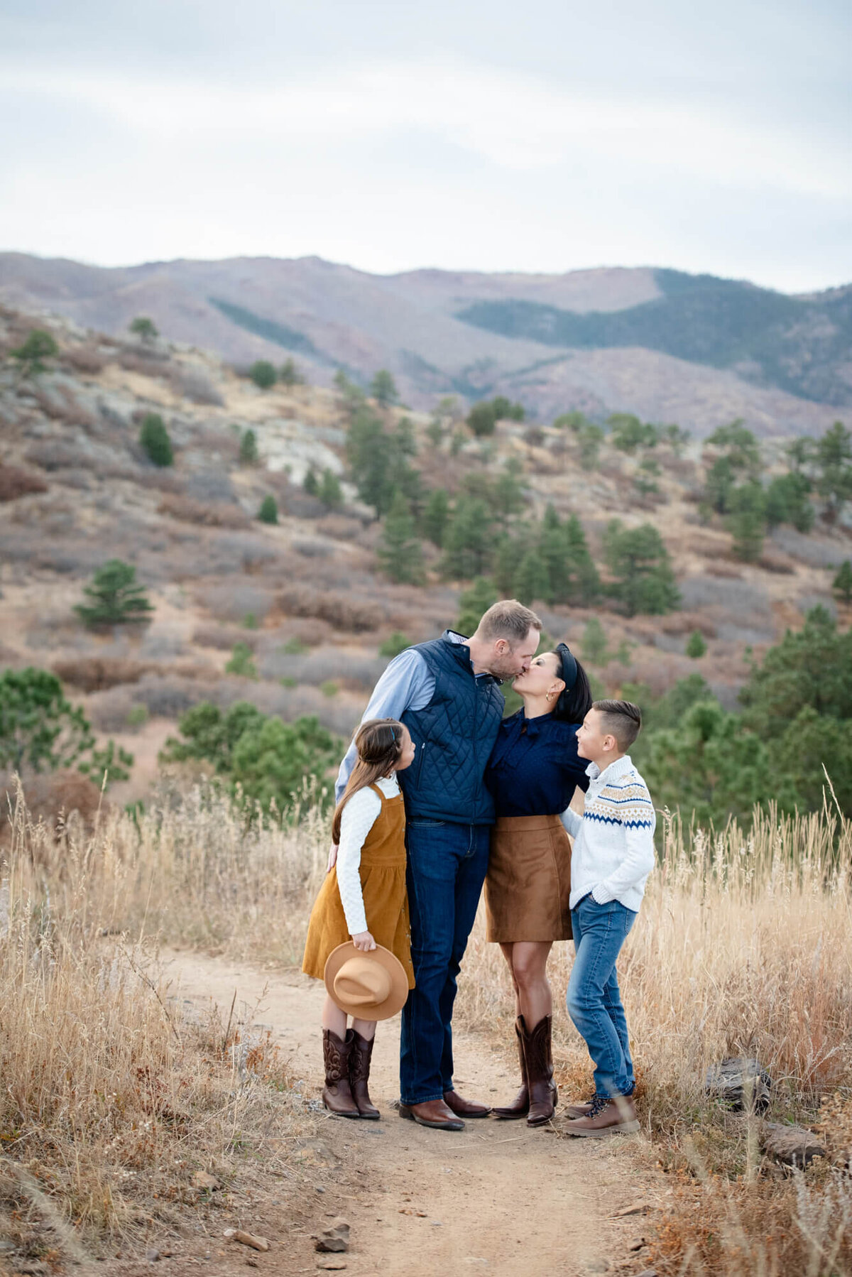 Mom and dad kiss while standing in a trail with their young son and daughter looking on