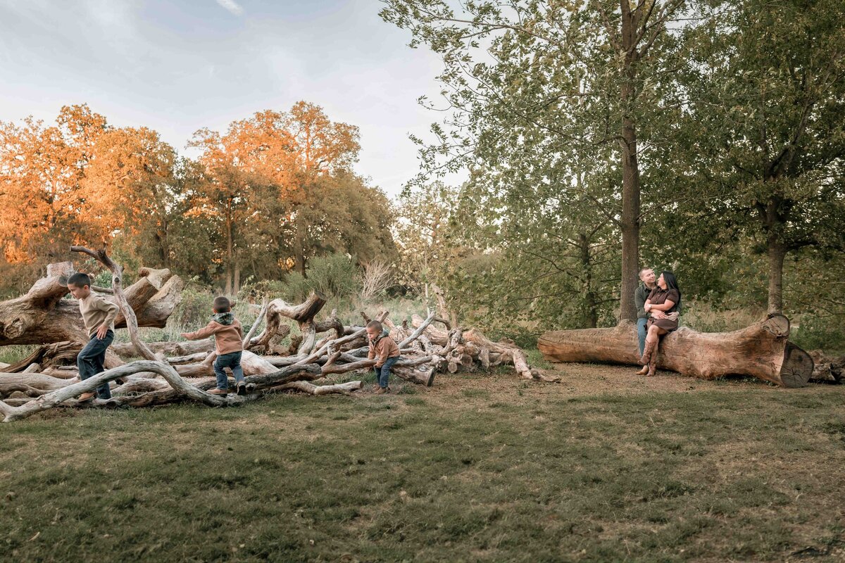 mom and dad cuddle each other while sitting on a log and watching their sons play and climb on broken down branches in Houston.