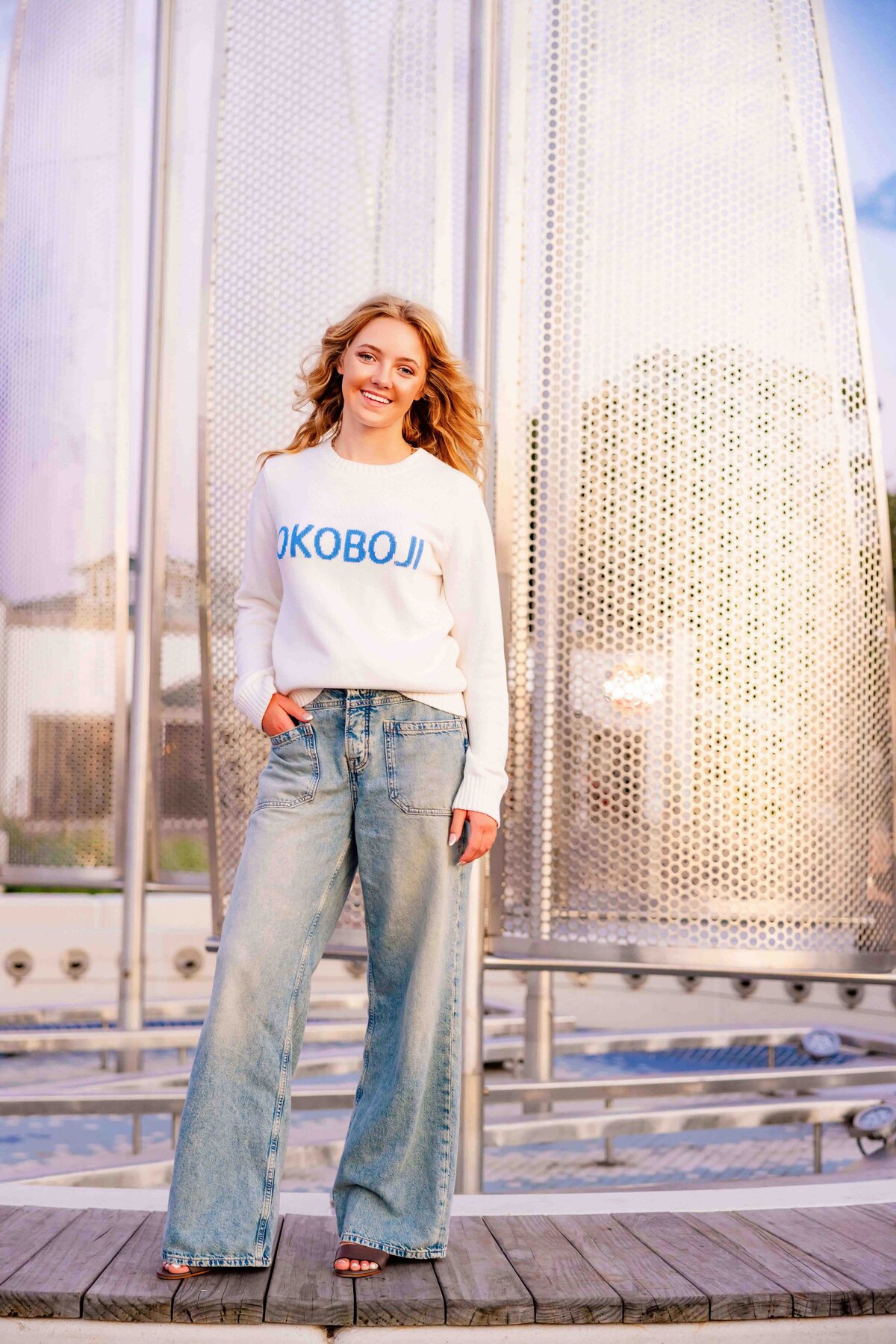 Senior girl in Okoboji sweatshirt standing in front of a metallic structure, casual and confident.