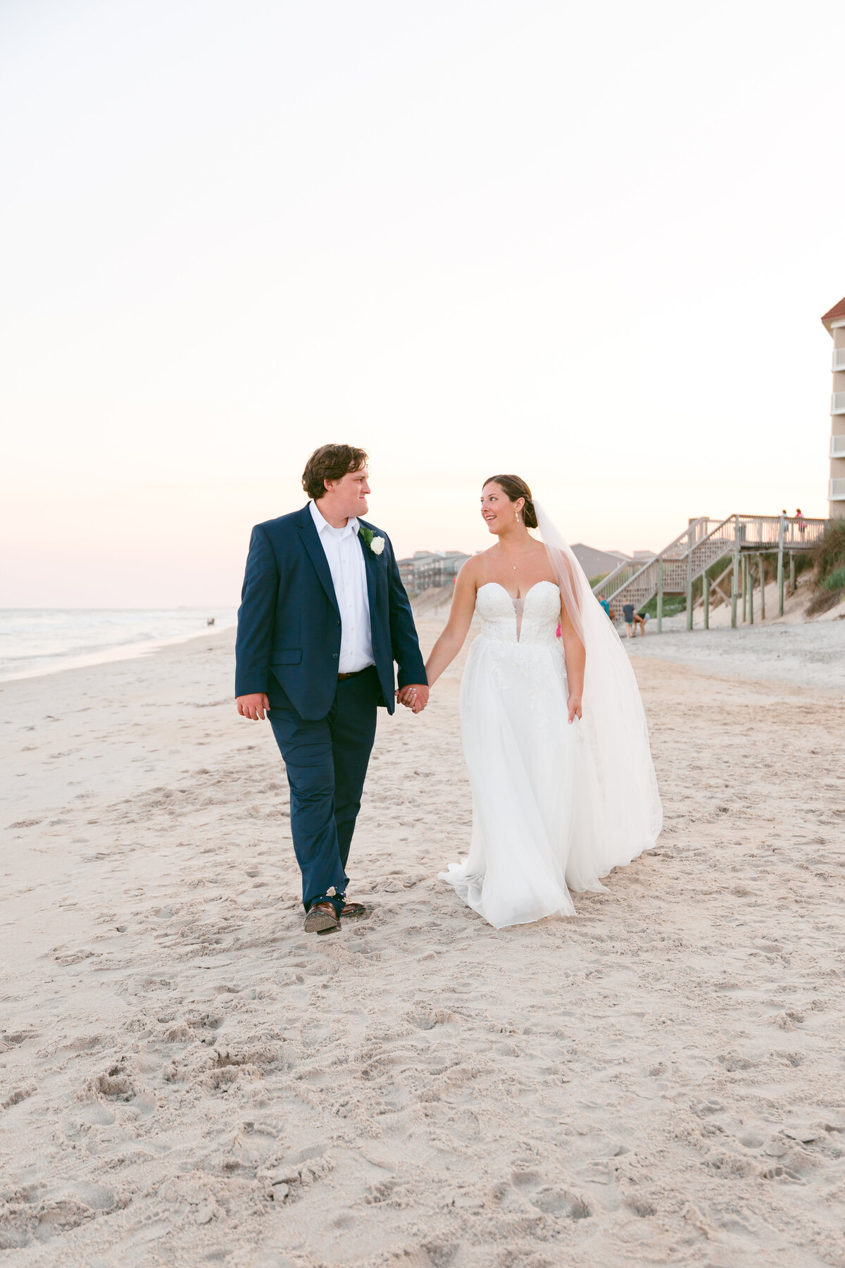 A couple in wedding attire walks barefoot along a sandy beach at sunset, holding hands, with houses visible in the distance.