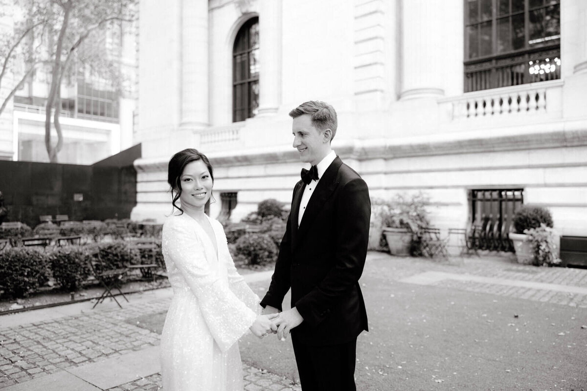 The groom is gazing at the bride while the latter is smiling towards the camera, in front of the NYPL, NYC. Image by Jenny Fu Studio