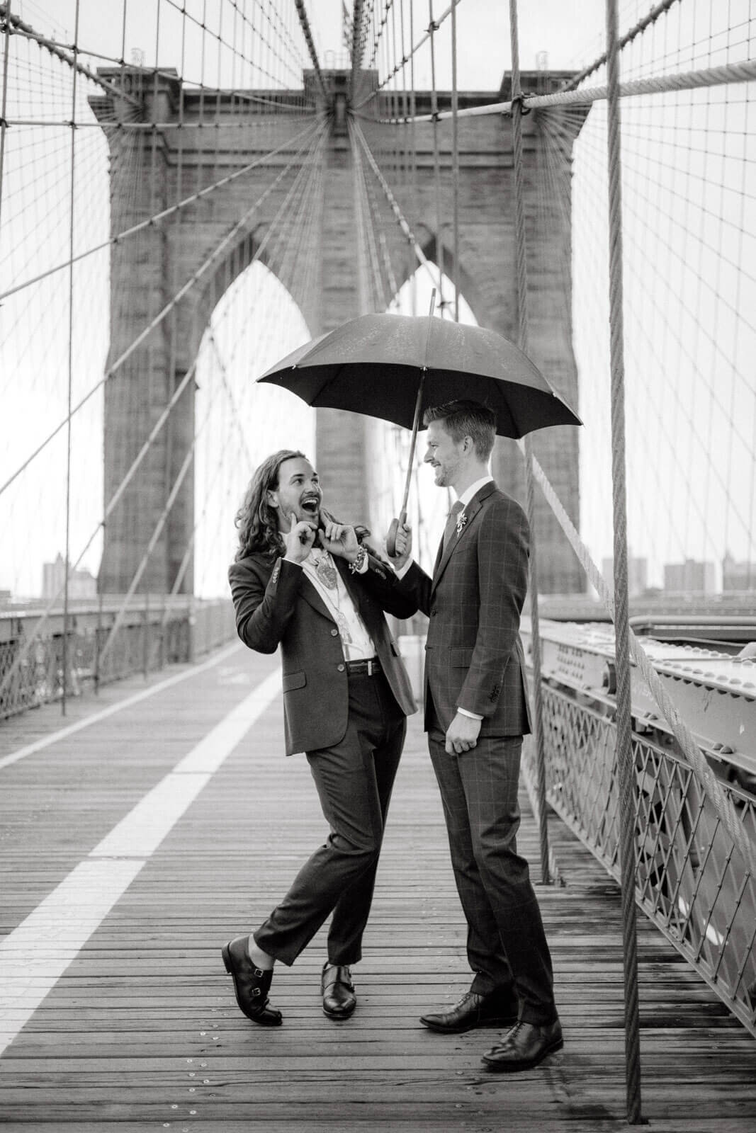 Black and white photo of the two grooms chatting under an umbrella on the Brooklyn Bridge. Elopement Image by Jenny Fu Studio