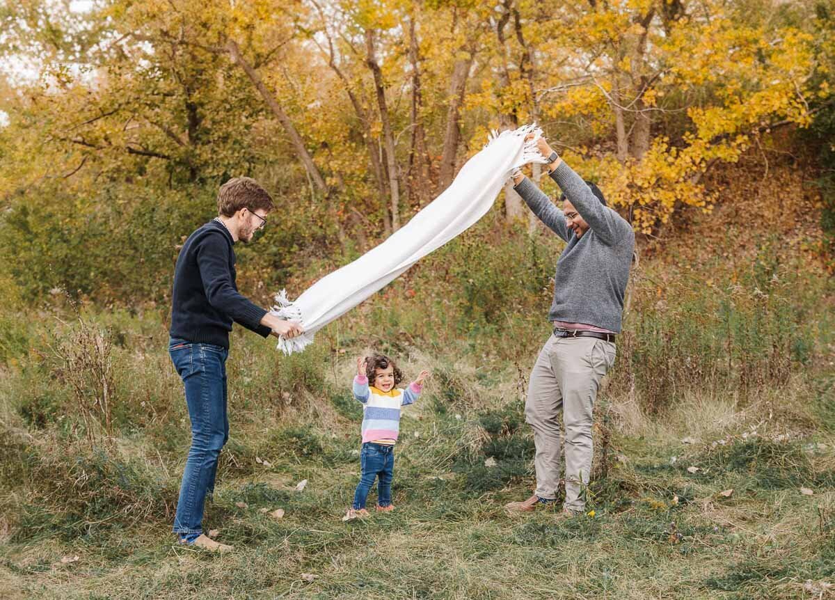 Young girl runs under a white blanket held up by her two dads. Playful moments captured by Toronto Family Photographer