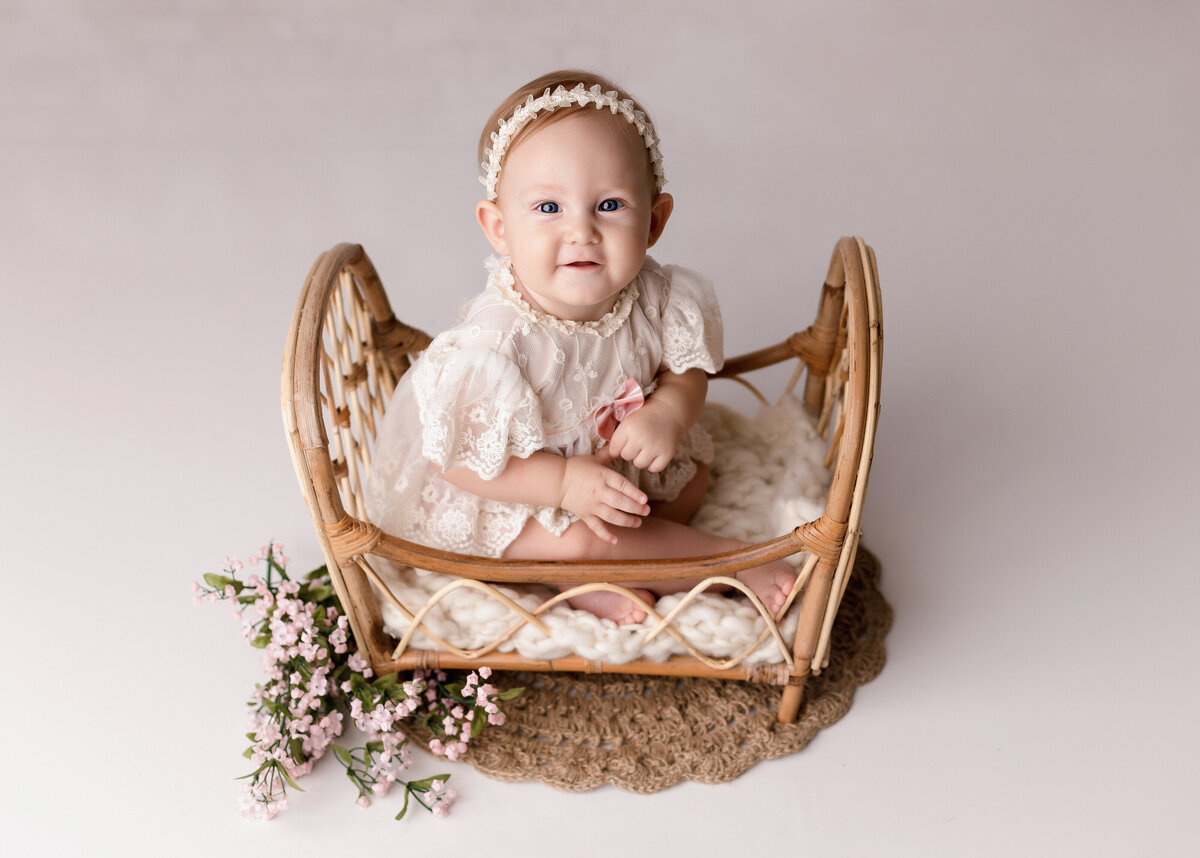 Baby girl 6-month milestone photoshoot at West Palm Beach and Wellington baby photo studio. Baby is wearing a vintage-inspired lace dress in a miniature rattan crib. Baby is looking up at the camera smiling.
