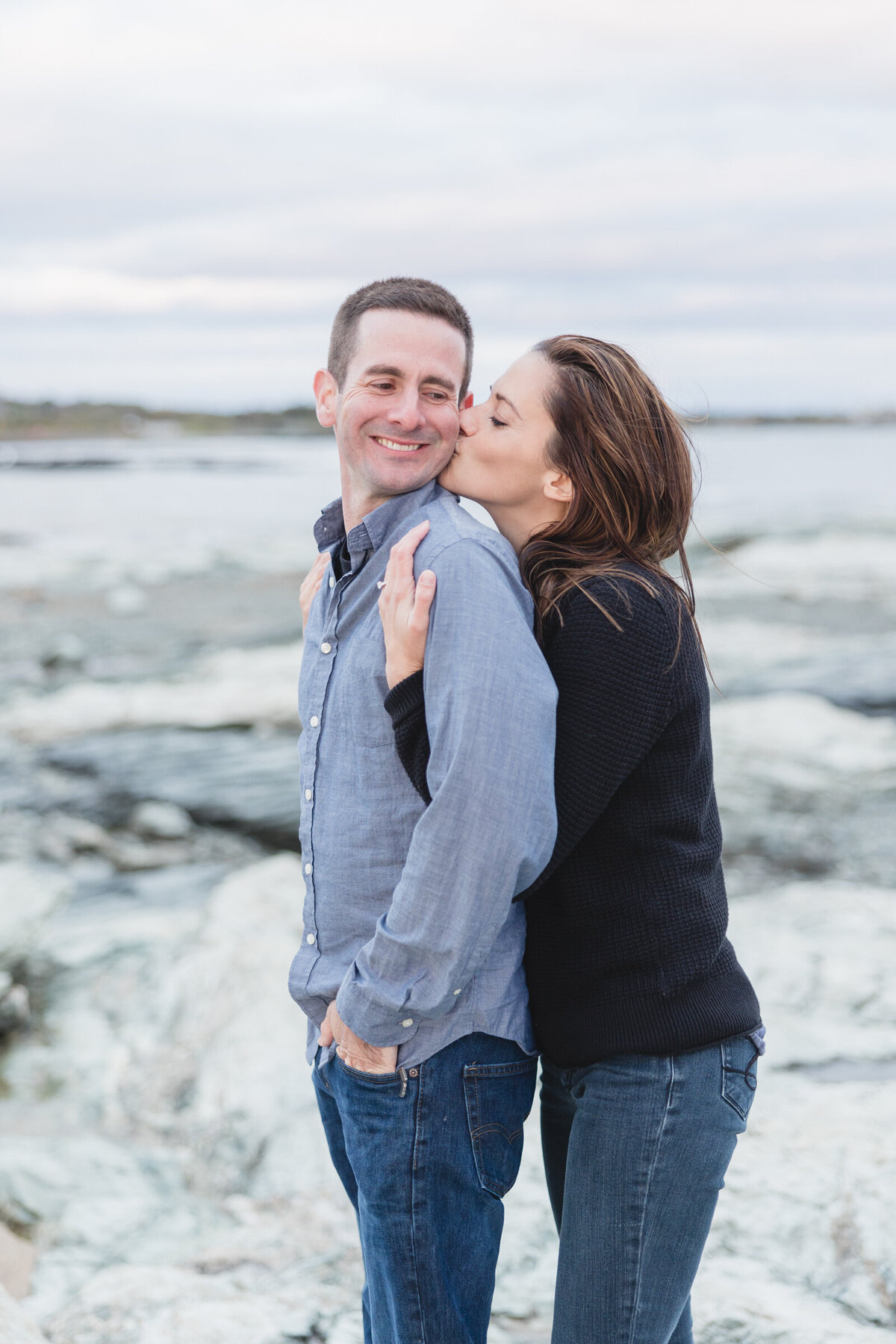 Woman kisses her fiancee on teh cheek during their engagement session in Newport